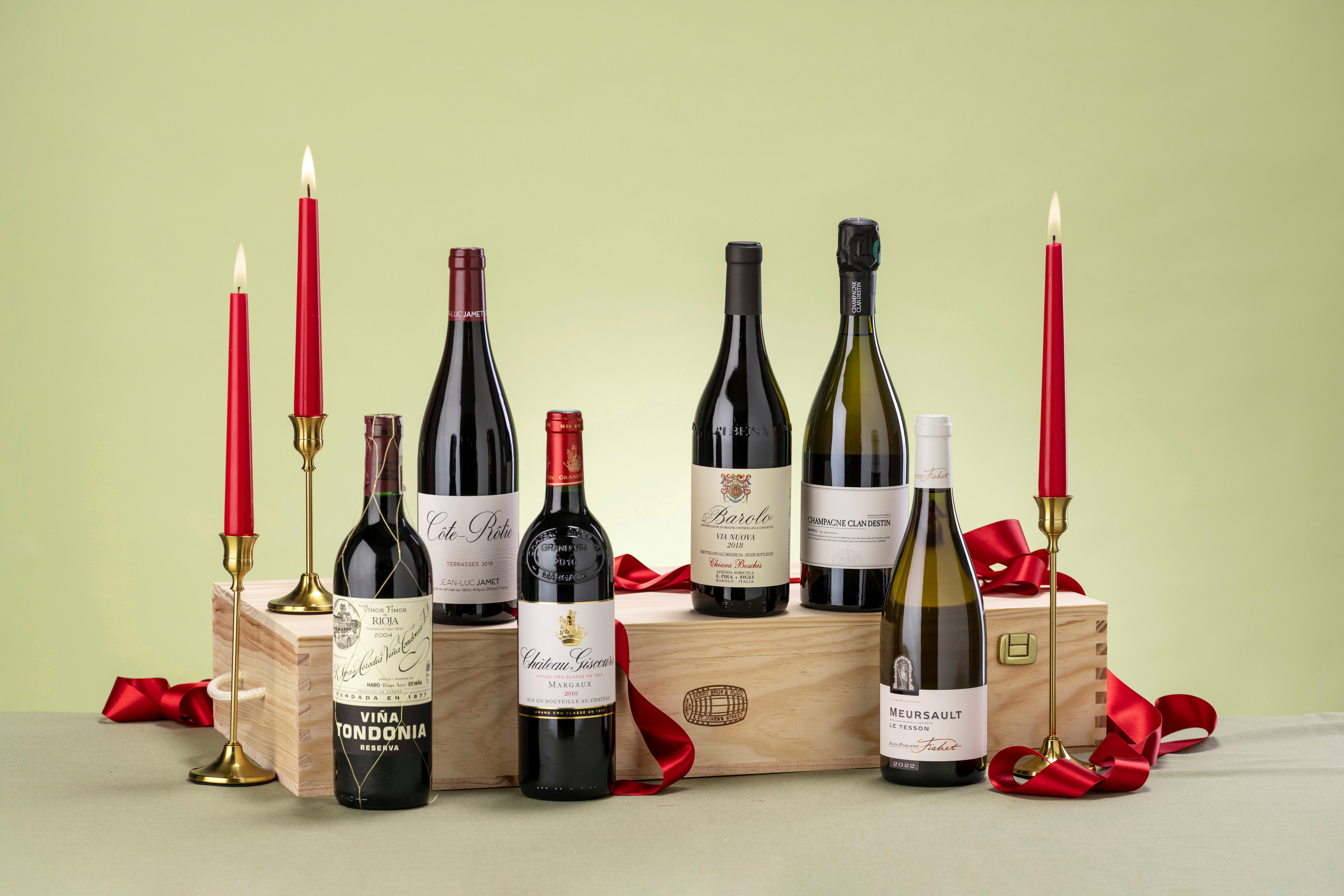 Six bottles of fine wine displayed on a wooden gift crate, surrounded by red ribbon and tall red candles, set against a light green background for a festive Christmas presentation.