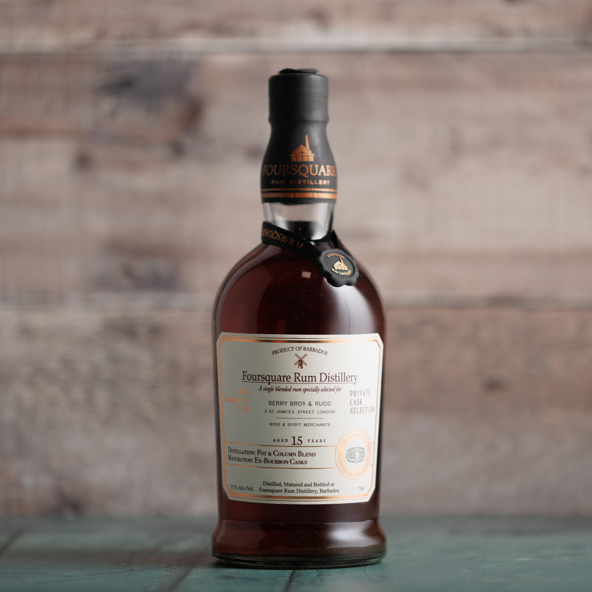 A photograph of Foursquare Distillery's 15-Year-Old Rum. The liquid is a deep ochre brown with gleaming hints of bronze on the label, and the bottle is standing against a faded wood-panelled wall