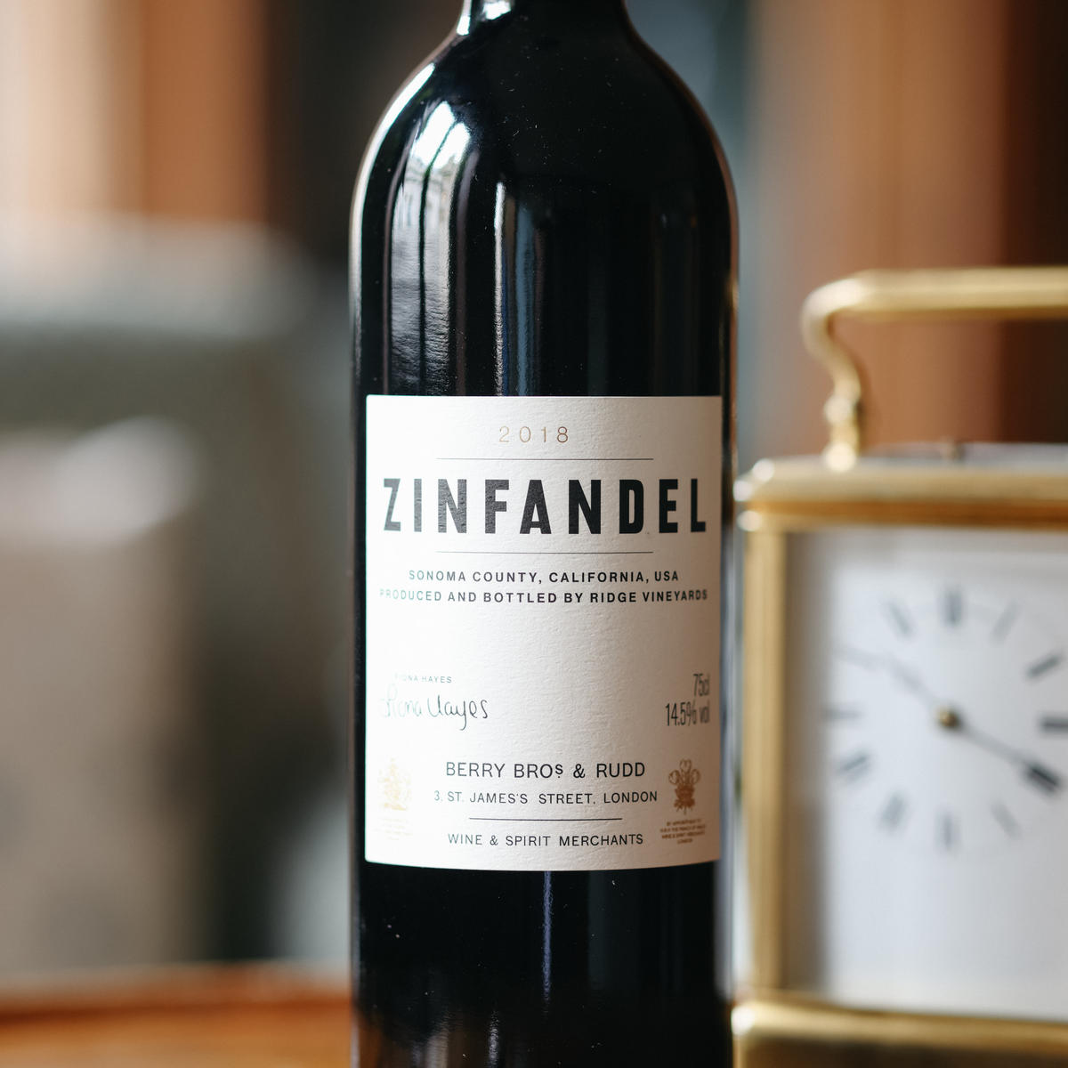 A photograph of our 2018 Own Selection Zinfandel standing on top of a table, with an old fashioned clock in the background