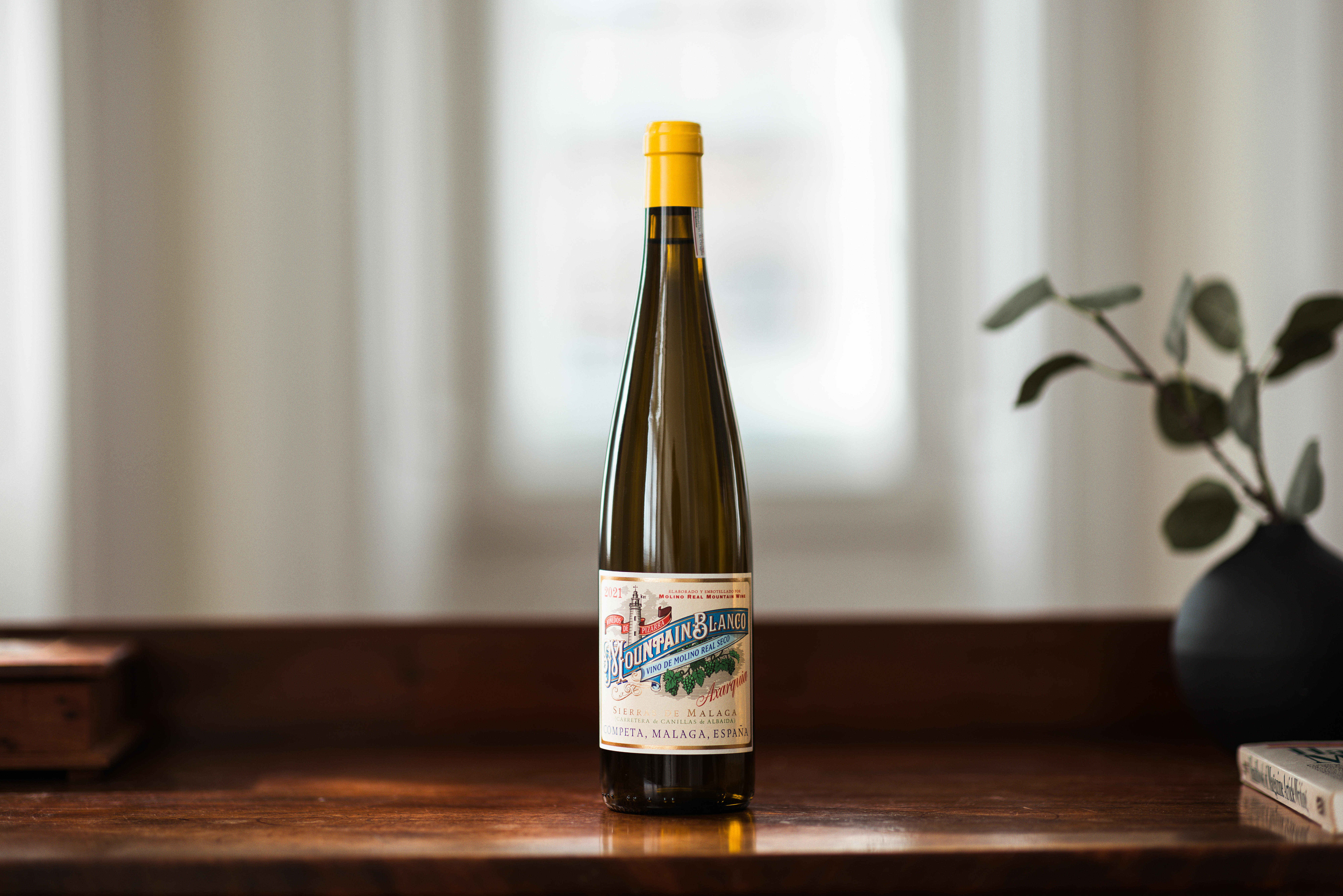 A bottle of Molino Real Mountain Blanco white wine with a yellow capsule, placed on a wooden surface in front of a softly blurred background.