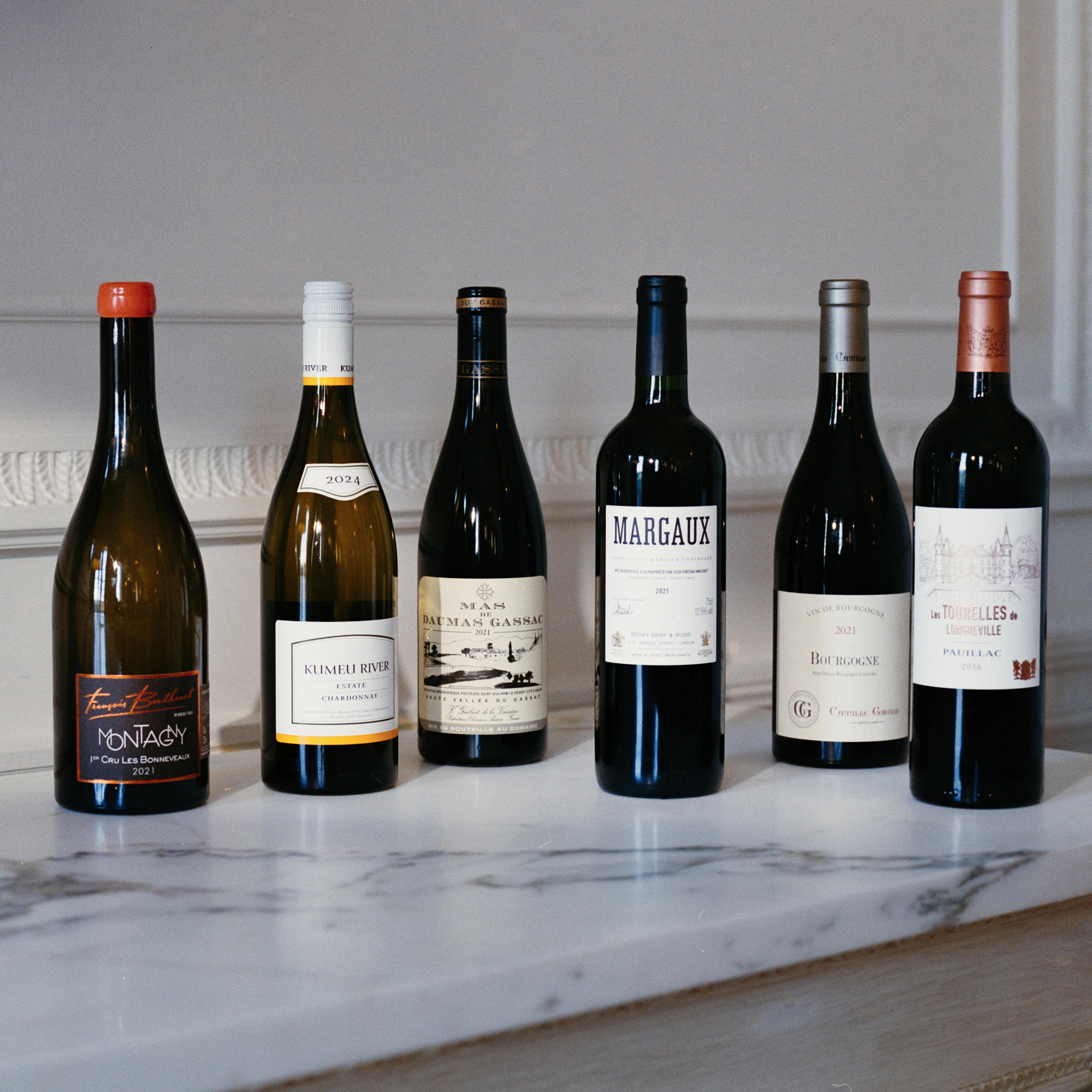  Six wine bottles, including red and white varieties, displayed side by side on a marble surface against a pale interior backdrop.