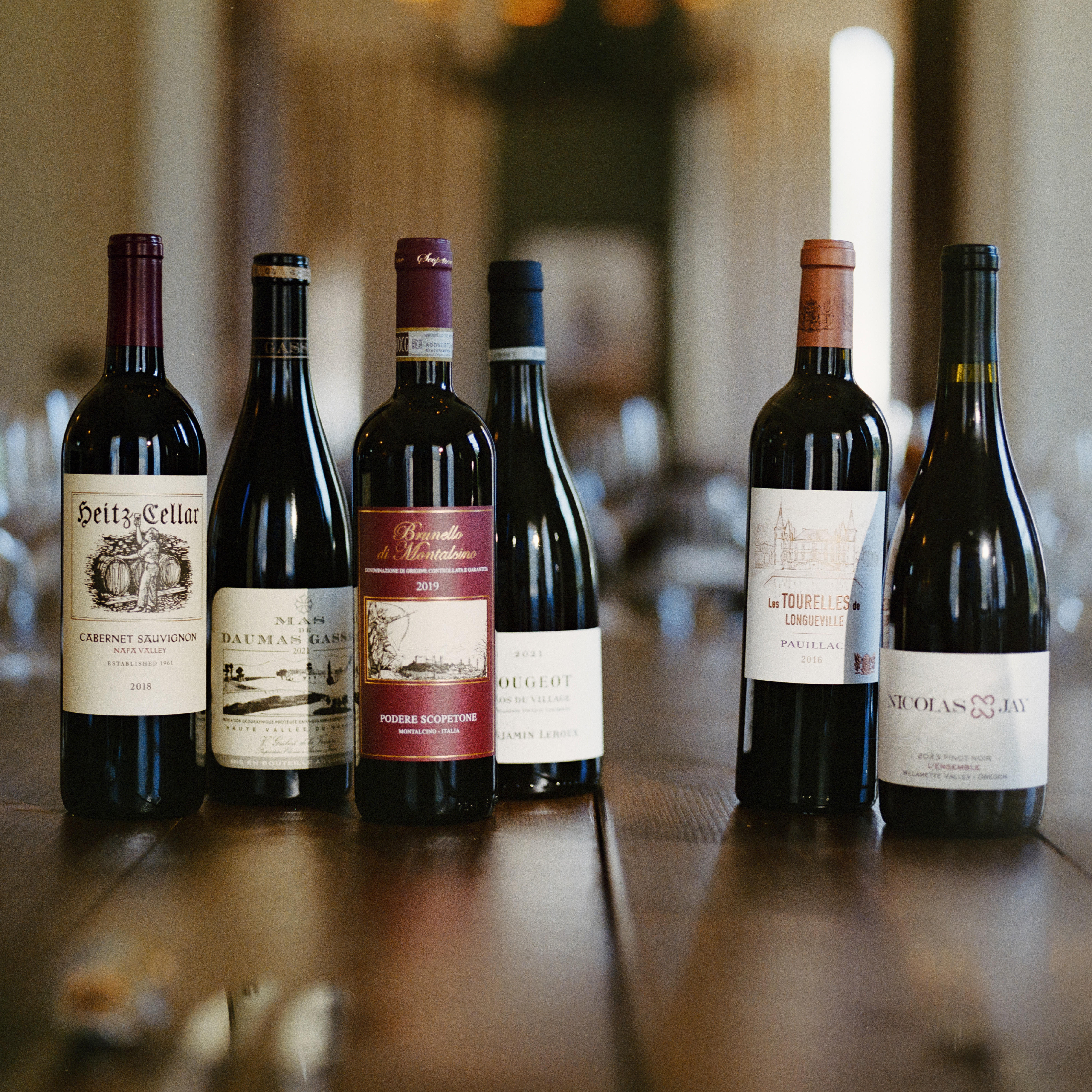 Six wine bottles displayed on a wooden table in a softly lit restaurant or tasting room. The bottles include a mix of red and white wines from various wineries, with labels facing forward and a blurred indoor background behind them.