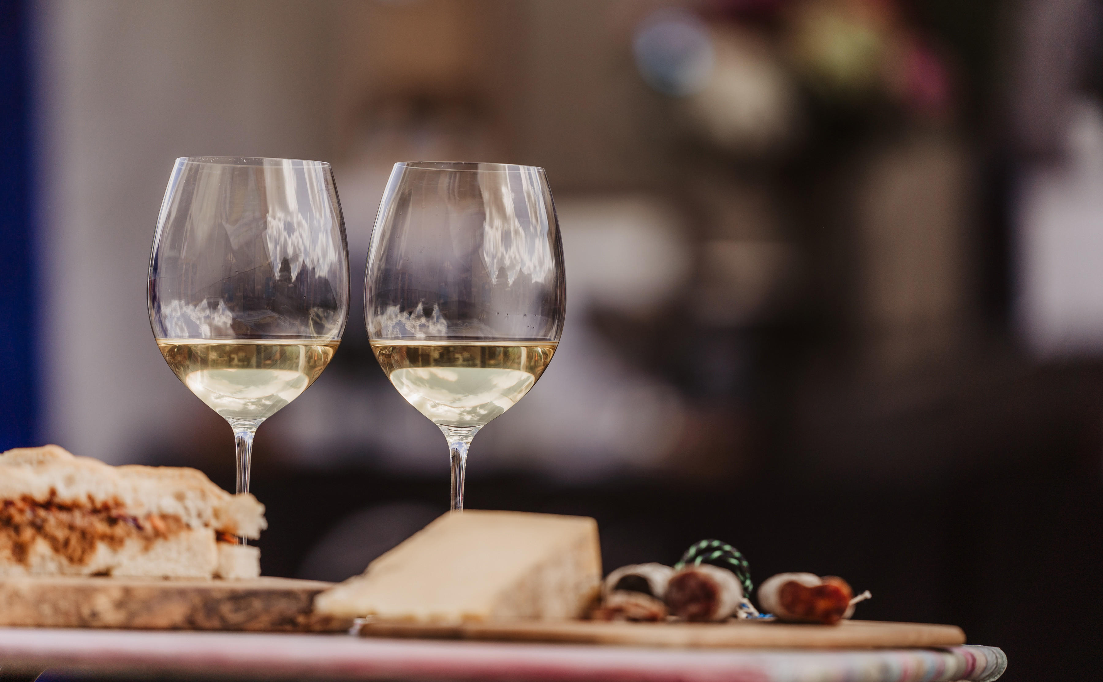 A photograph of two glasses of white Burgundy sitting atop a table beside a sandwich and a block of cheese