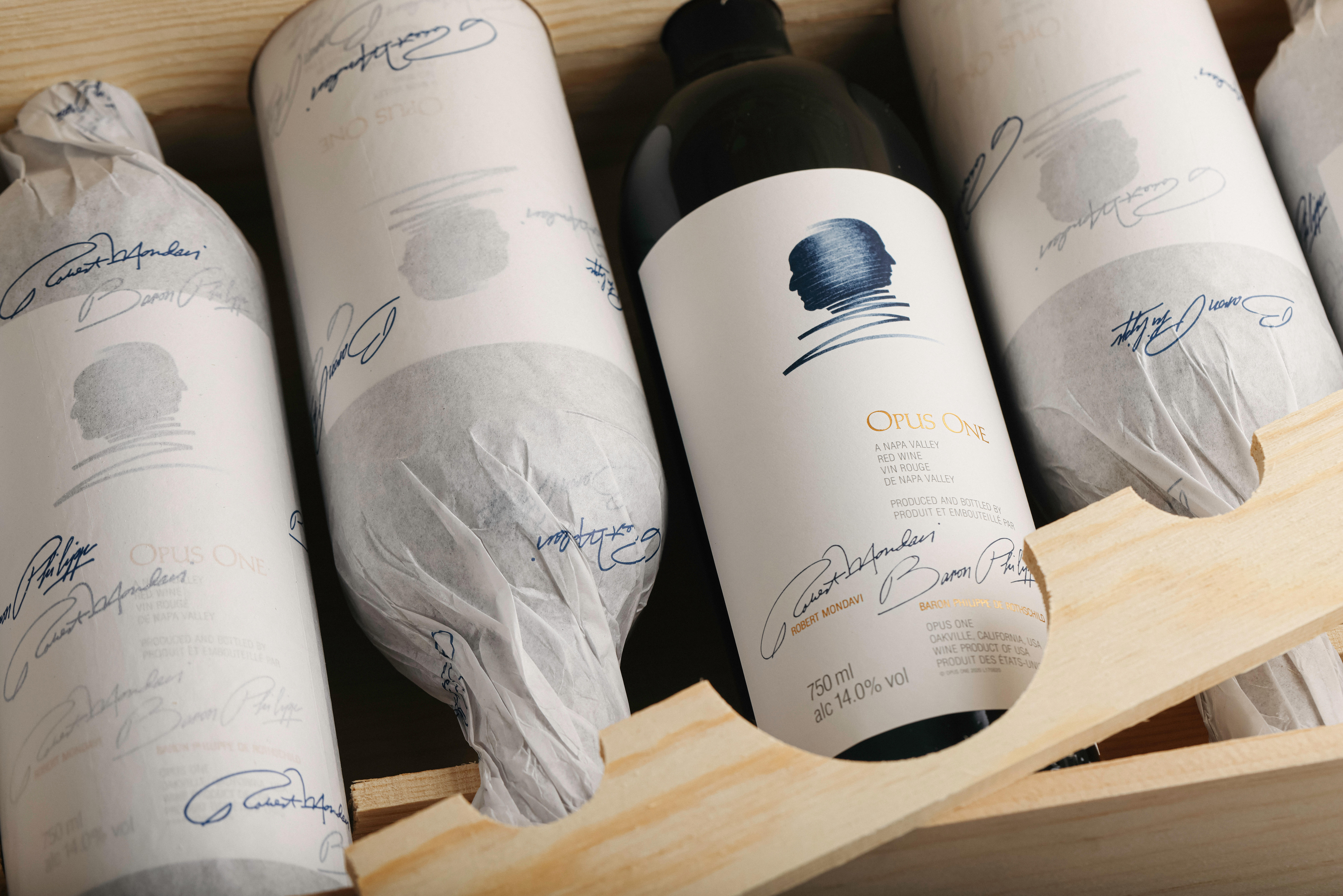 Opus One wine bottles in a wooden crate, with one bottle prominently displayed and others wrapped in white tissue paper.
