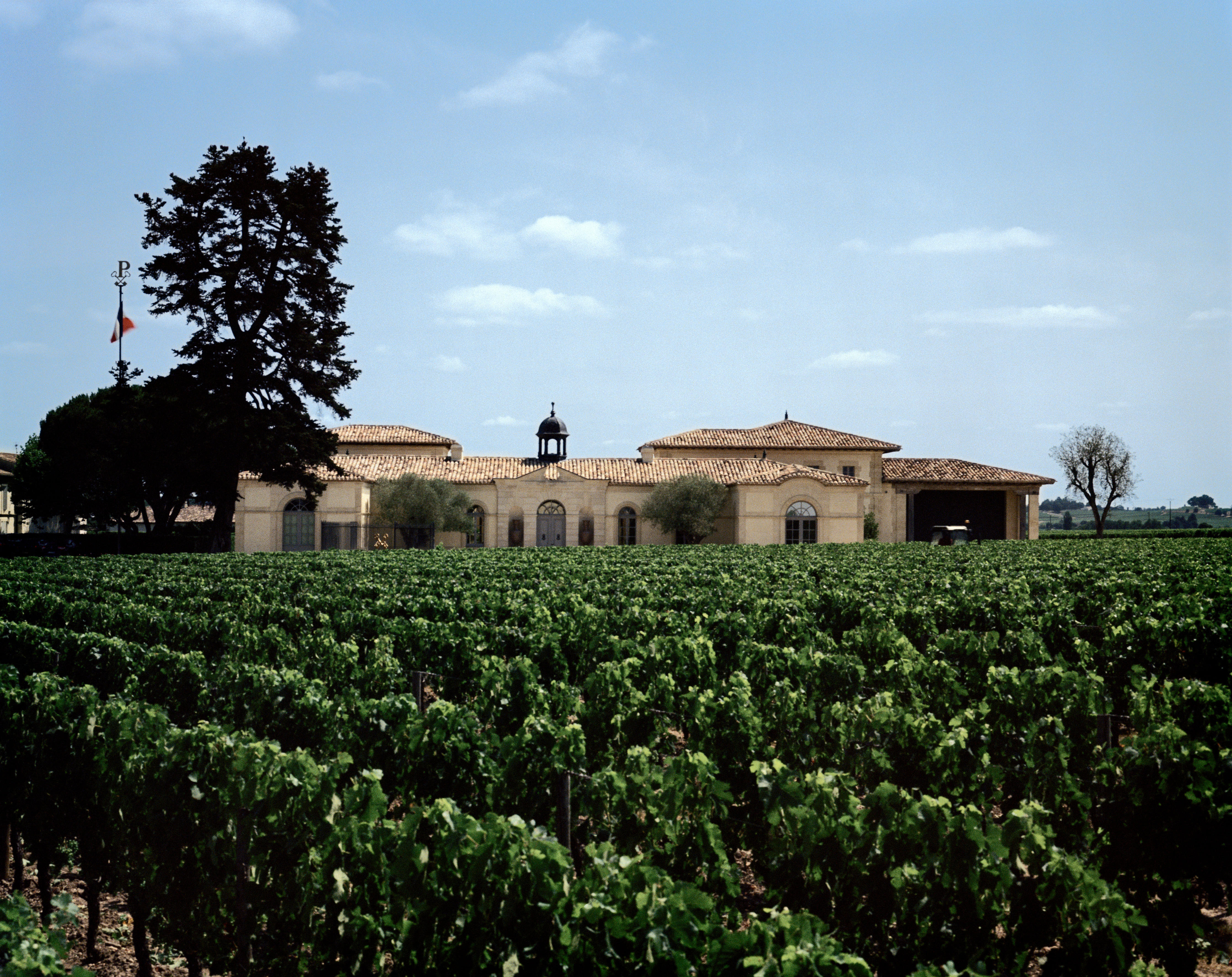 The Petrus estate in Pomerol, Bordeaux, featuring a grand stone building with a central cupola and terracotta roof tiles, surrounded by lush green vineyards under a clear blue sky.