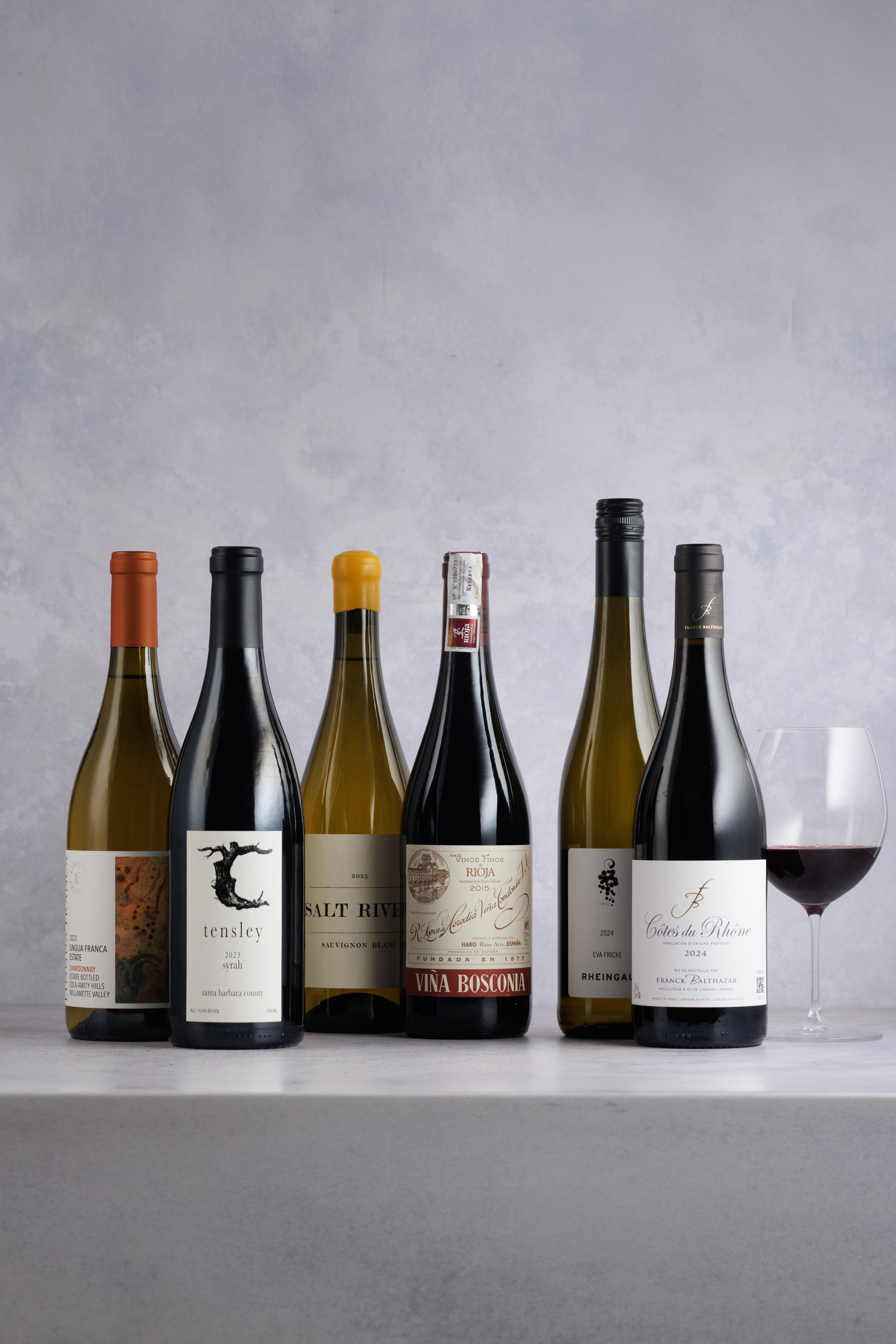 6 bottles of fine wines from regions like South Africa and Spain with a glass of red wine on the right side against a grey background.