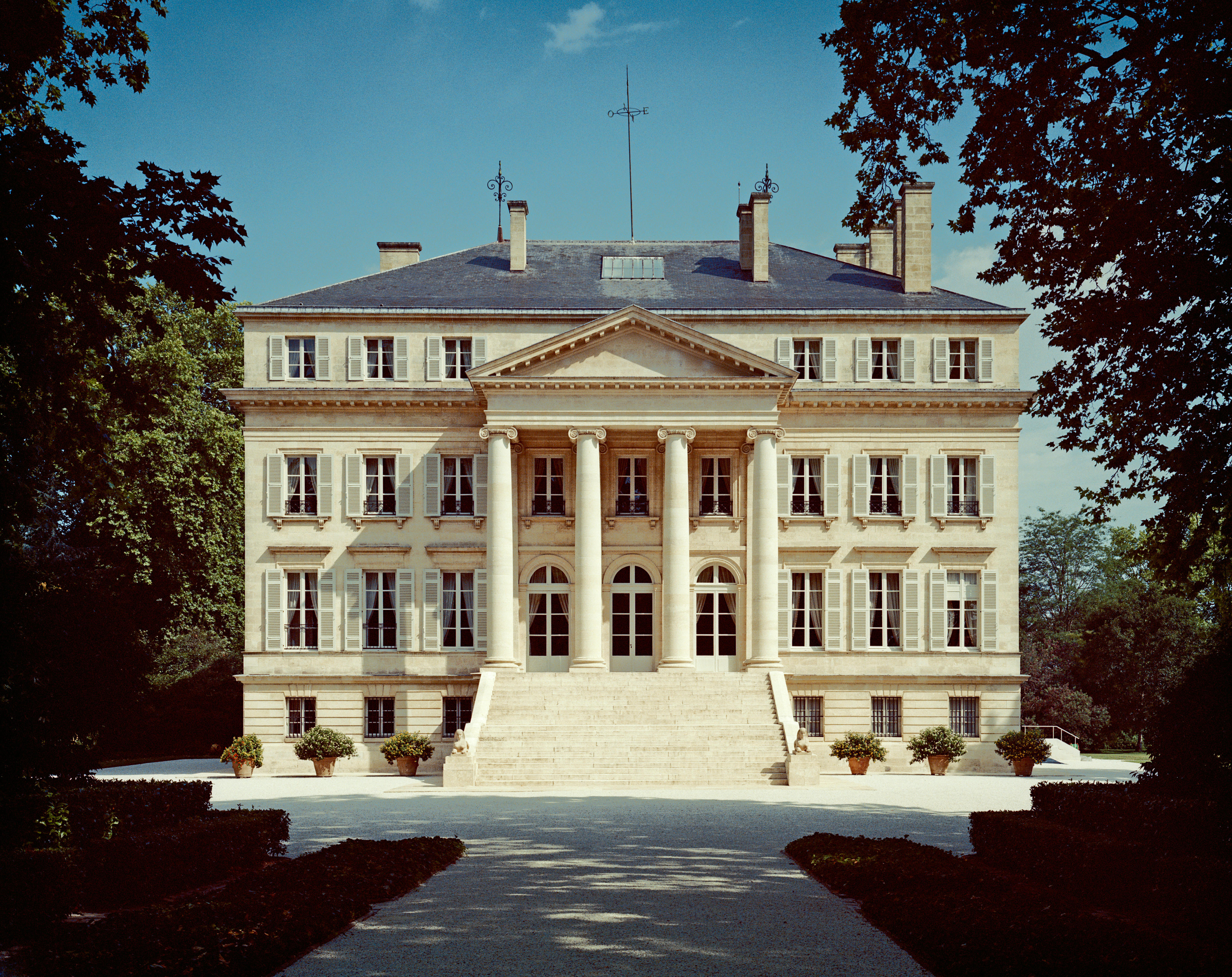 The neoclassical façade of Château Margaux in Bordeaux, featuring symmetrical architecture with tall columns, arched windows, and a grand staircase, framed by trees under a bright blue sky.