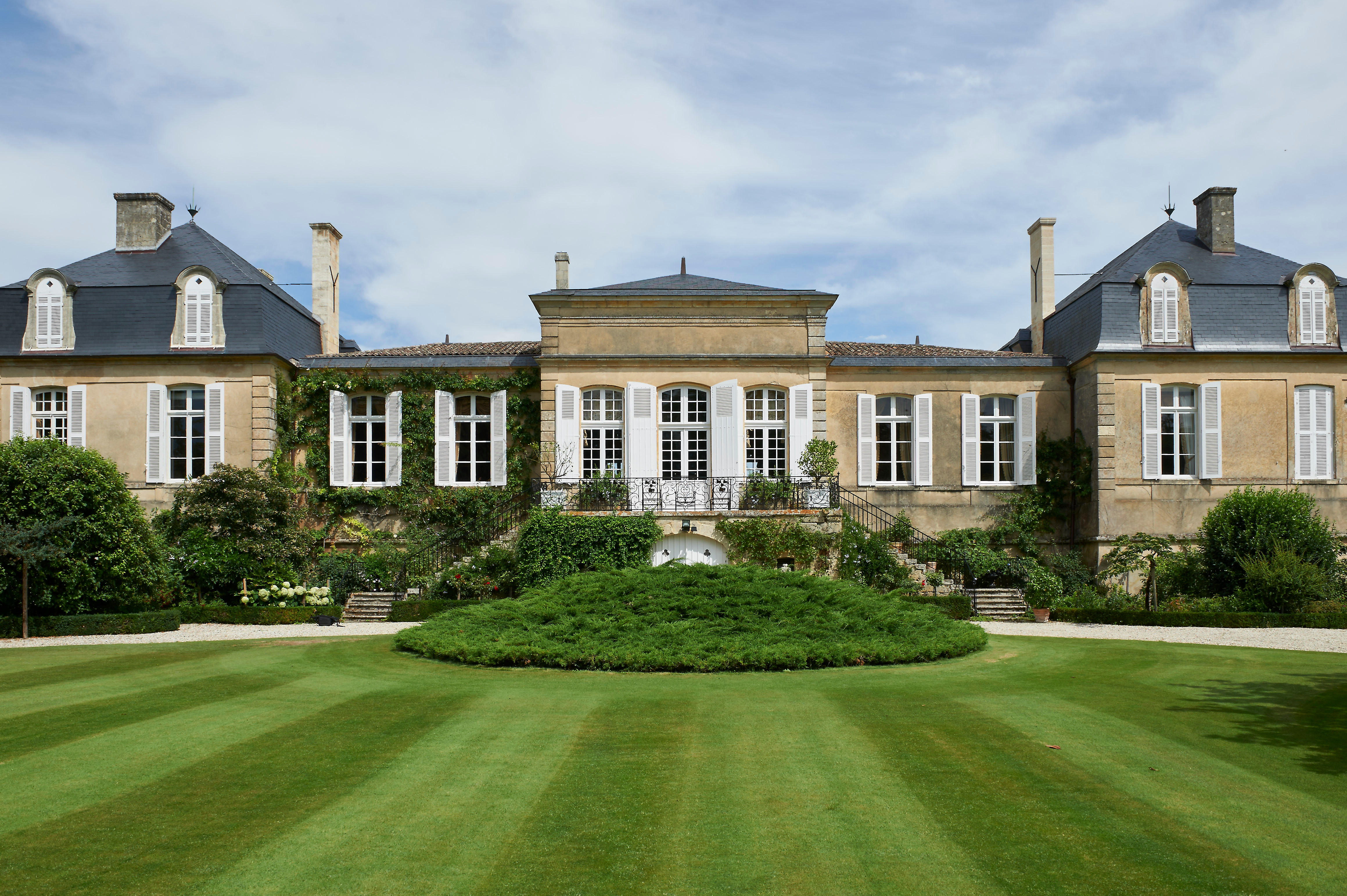 Front view of Château Langoa-Barton, a historic Bordeaux estate with symmetrical architecture, tall white-shuttered windows, ivy-covered walls, and a manicured circular lawn in the foreground under a partly cloudy sky.