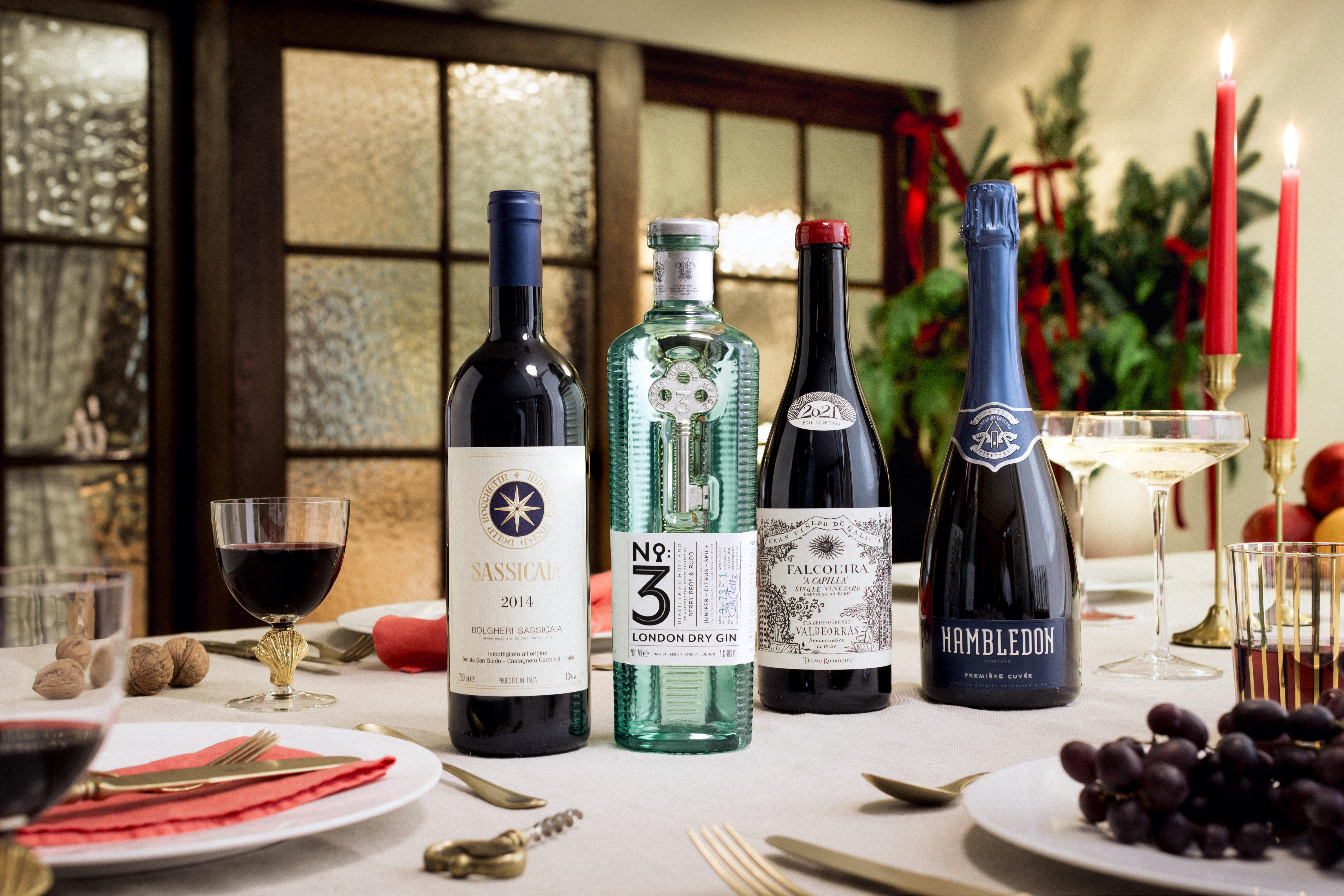 Festive dining table featuring a selection of wines and spirits: a bottle of Sassicaia 2014 red wine, No.3 London Dry Gin in a green glass bottle, Falcoeira red wine 2021, and Hambledon sparkling wine.