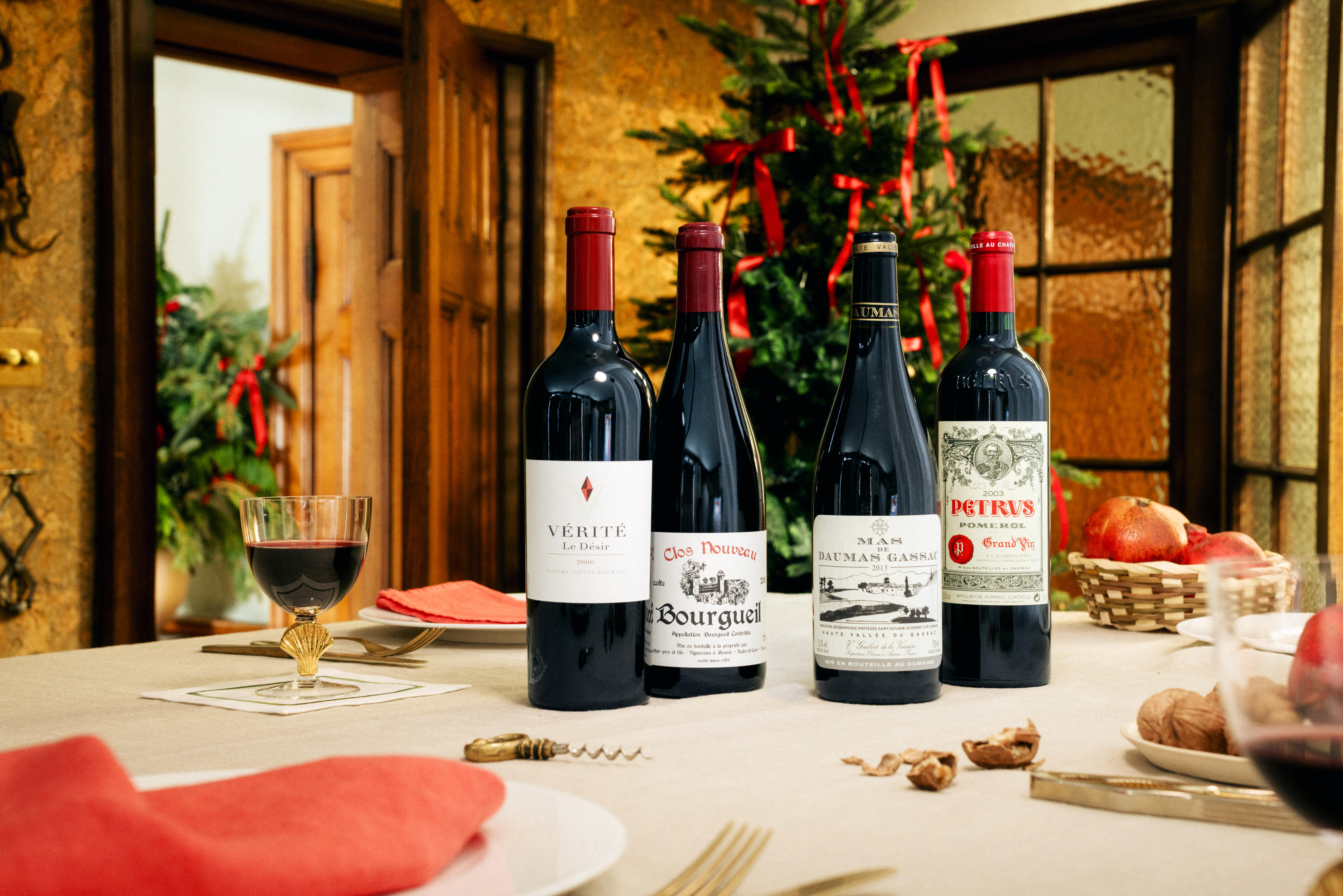 Four bottles of mature fine wine displayed on a festive dining table set for Christmas. The wines include Vérité La Désir, Clos Nouveau Bourgueil, Mas Daumas Gassac, and Petrus Pomerol. A glass of red wine sits to the left, with holiday decorations.