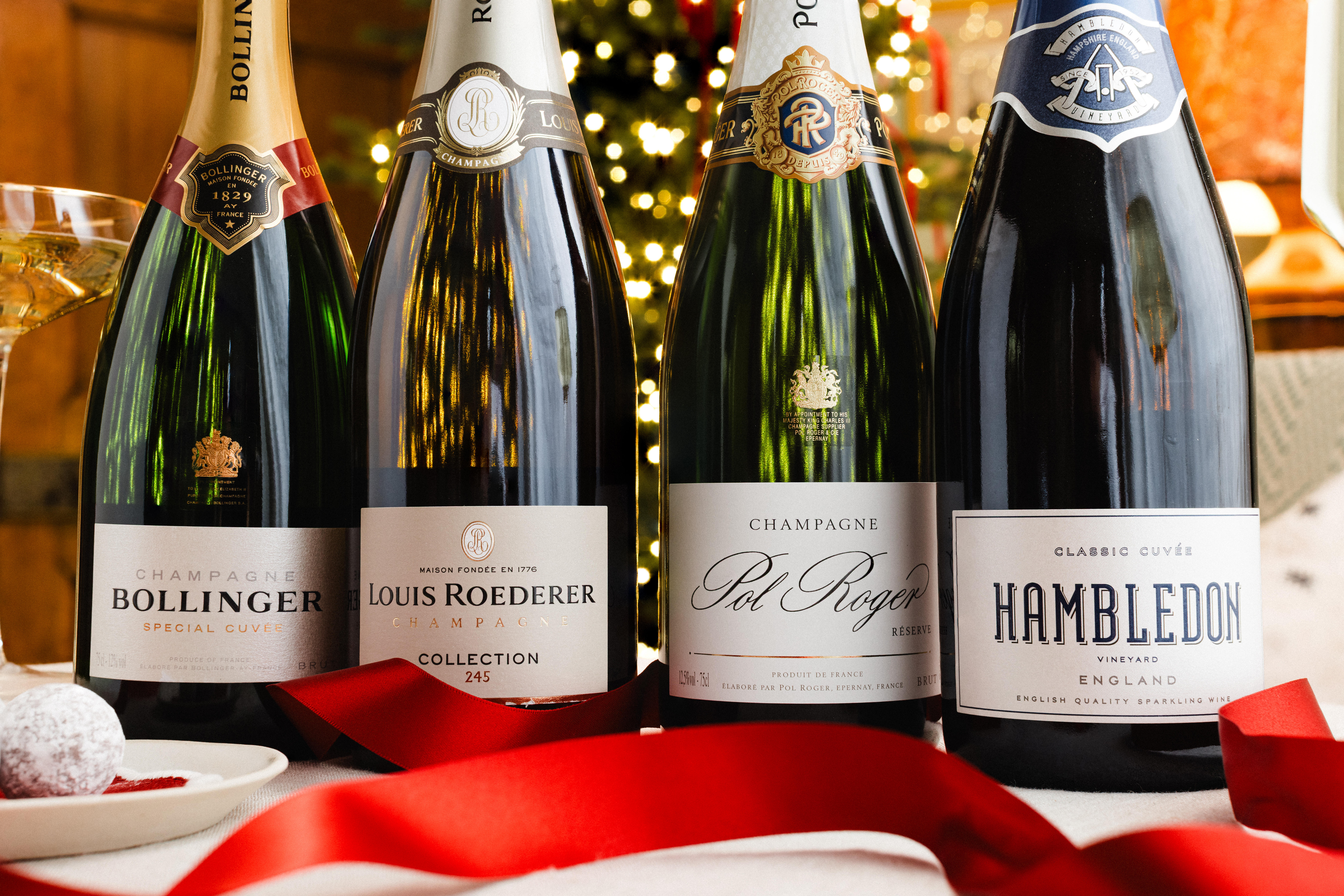 Four bottles of sparkling wine and Champagne displayed side by side: Bollinger Special Cuvée, Louis Roederer Collection 245, Pol Roger Réserve, and Hambledon Classic Cuvée. A red ribbon and festive decorations are placed in the foreground.