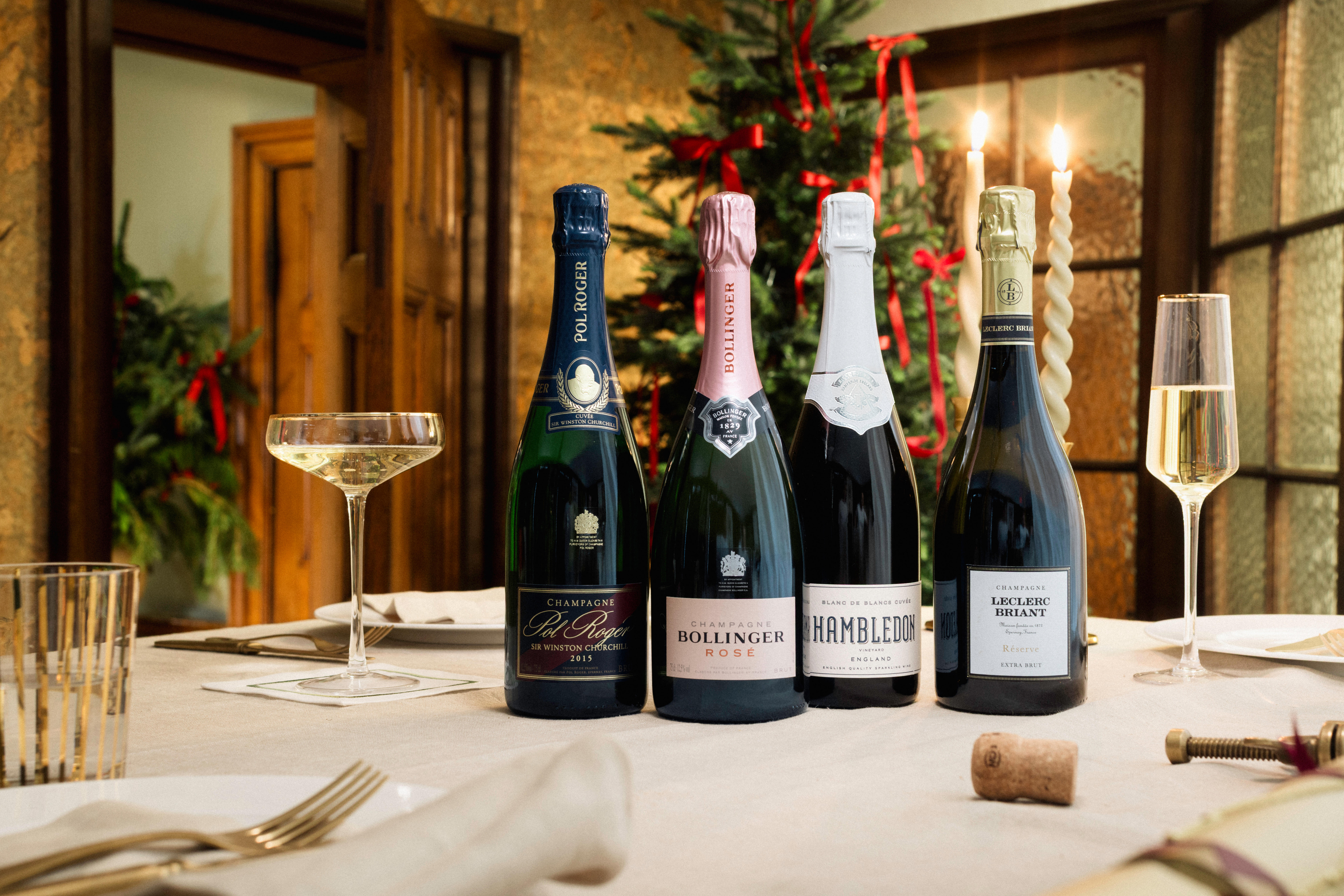 Four bottles of Champagne and sparkling wine on a festive table with Christmas décor, including Pol Roger, Bollinger Rosé, Hambledon, and Leclerc Briant.
