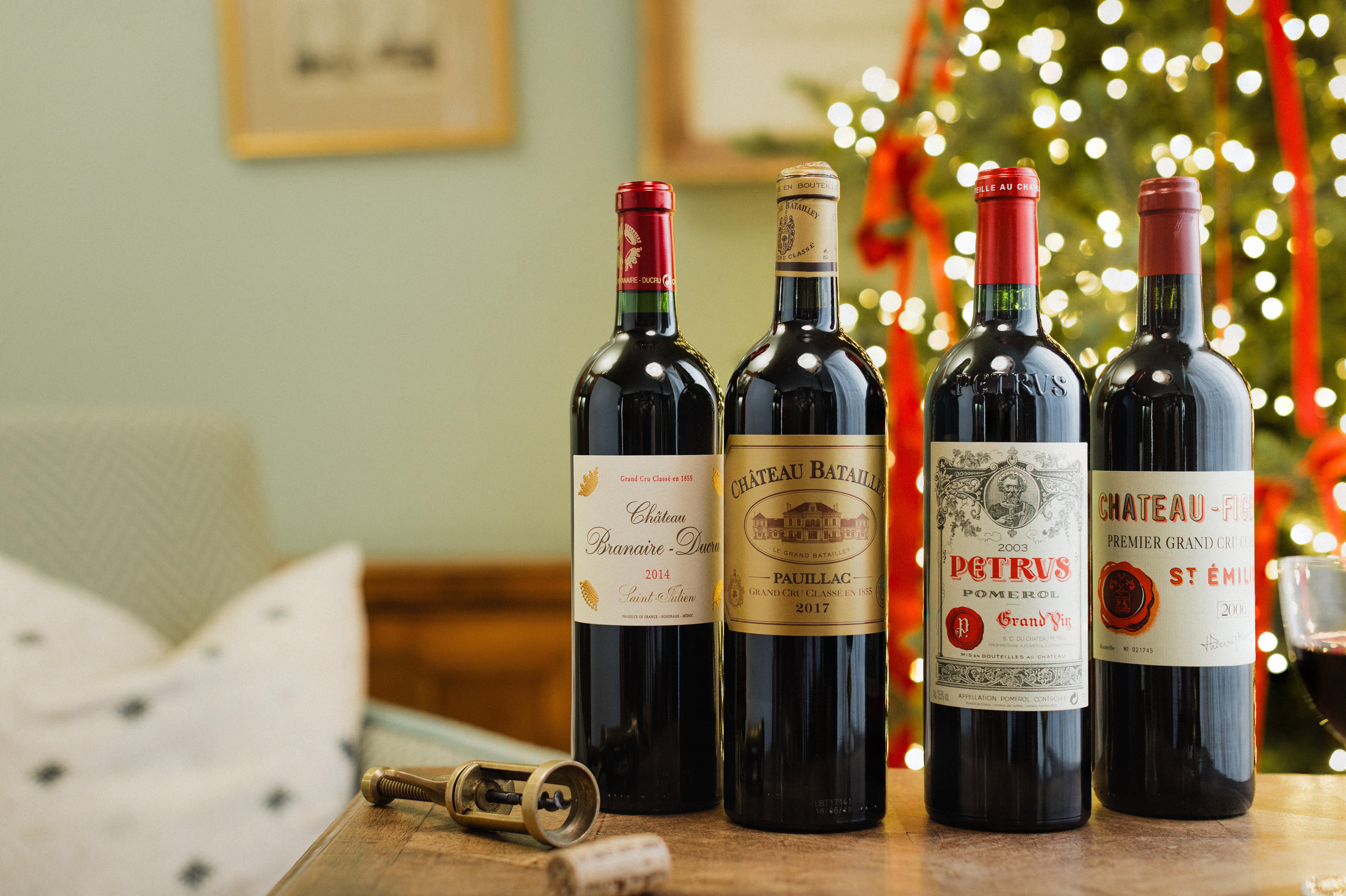 Four bottles of fine Bordeaux red wine displayed in a festive setting: Château Branaire-Ducru 2014, Château Batailley 2017, Petrus Pomerol 2003, and Château-Figeac Premier Grand Cru Classé from Saint-Émilion.
