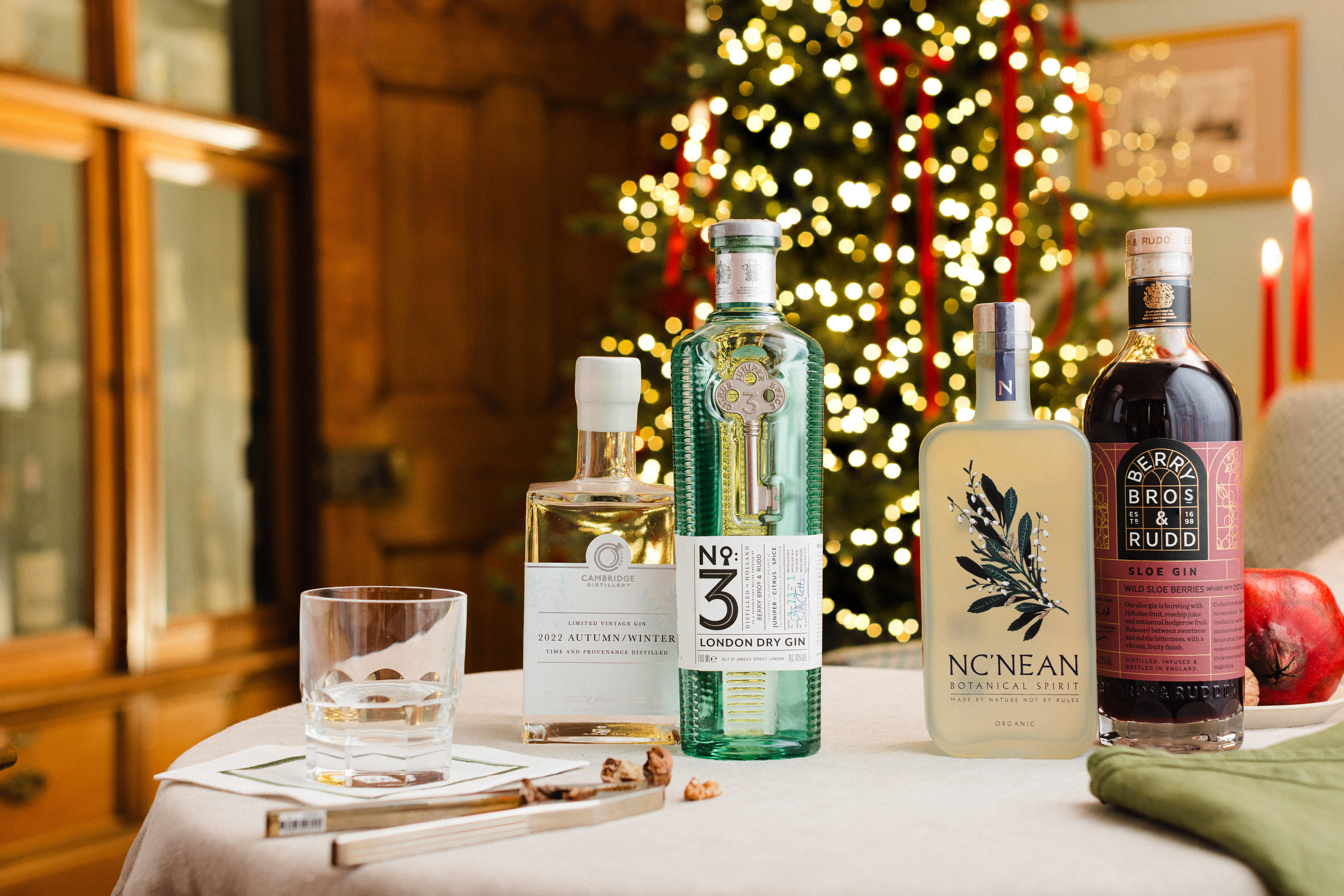 Four bottles of premium gin displayed on a festive table with a glass and garnishes, set against a background of a decorated Christmas tree and glowing lights.