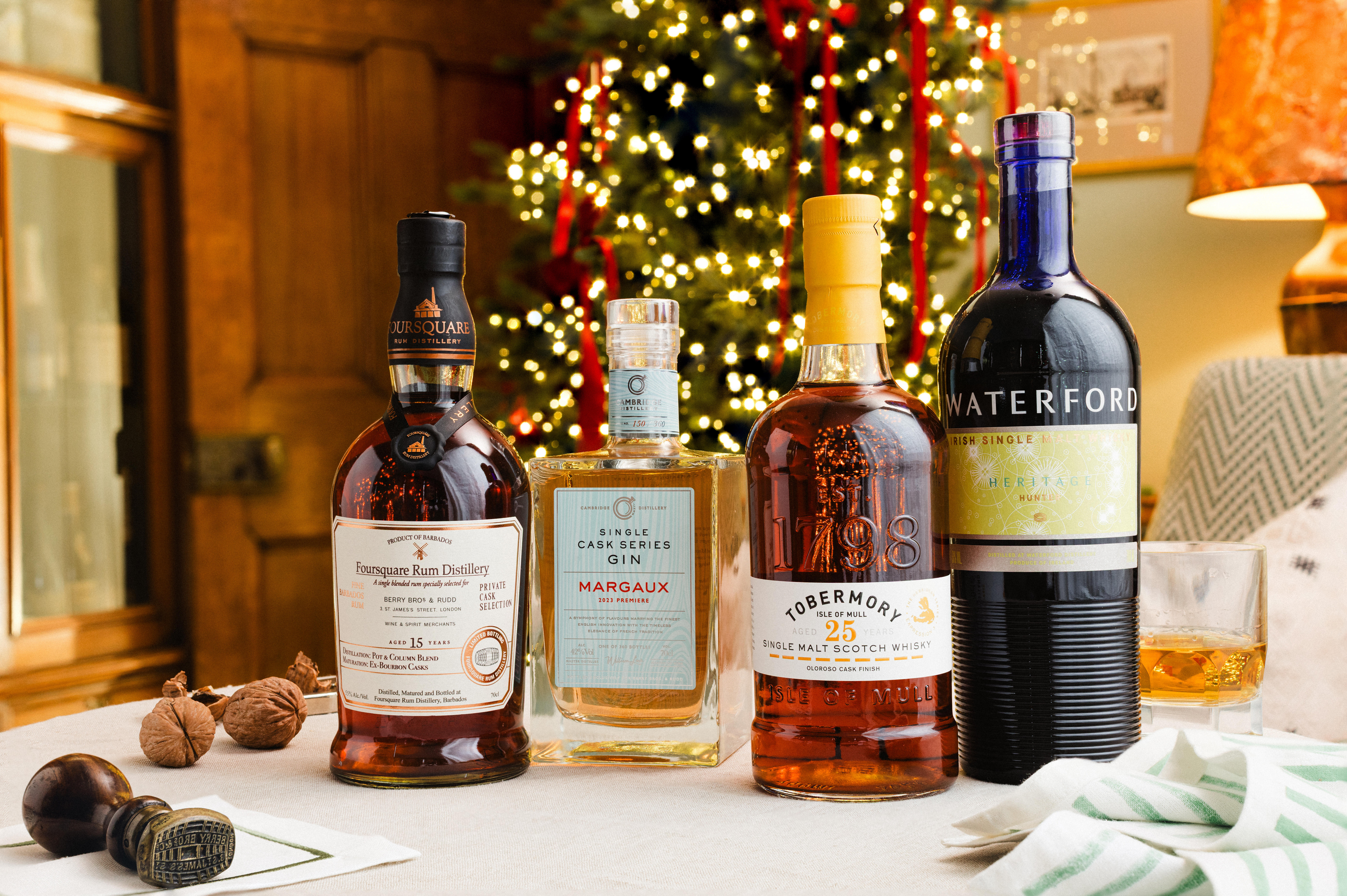A festive table featuring four bottles of fine spirits: Foursquare Rum, Margaux Cask Series Gin, Tobermory 25-Year-Old Whisky, and Waterford Single Malt, set in front of a Christmas tree with glowing lights and red ribbons.