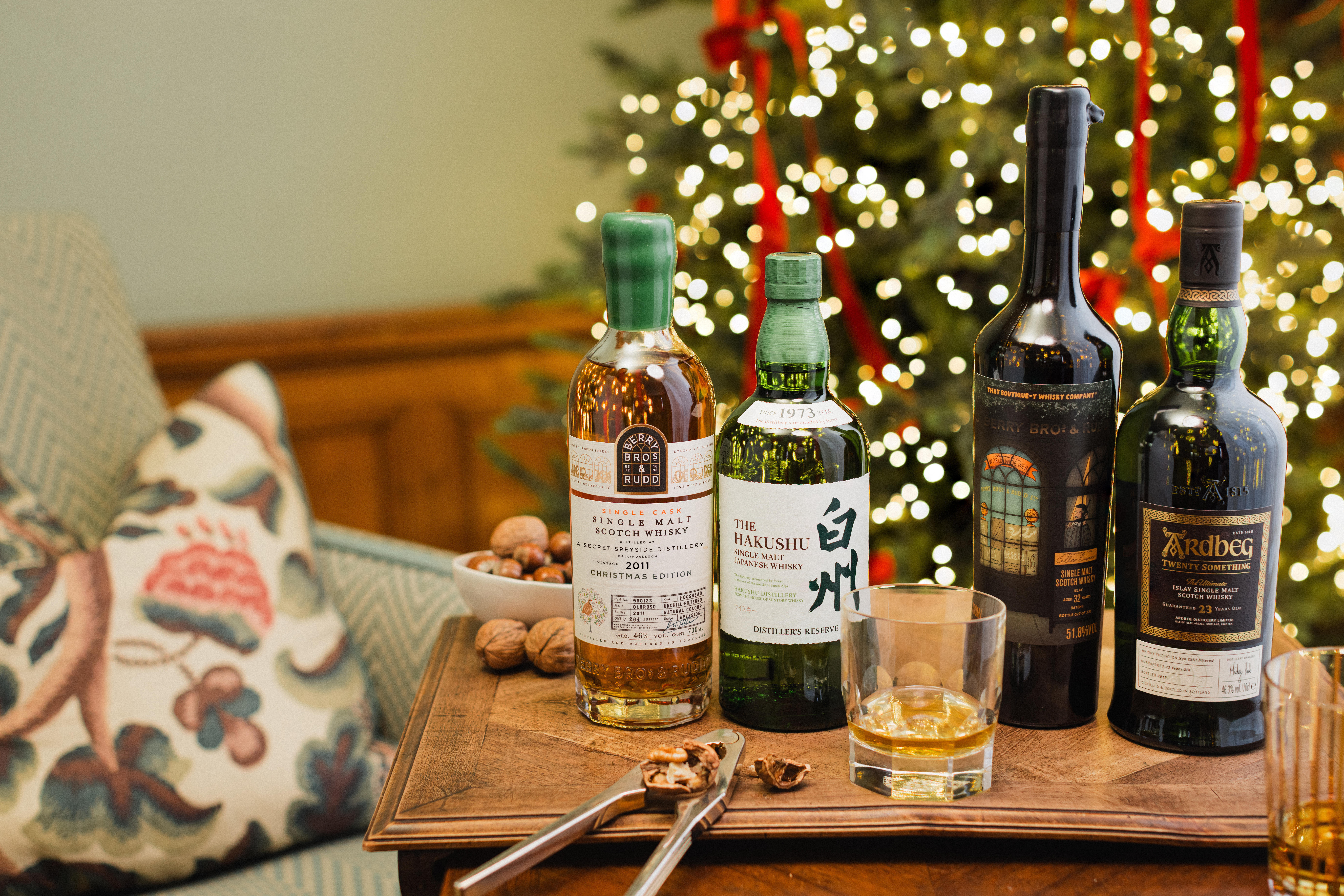 A wooden tray displaying four bottles of premium whisky in front of a Christmas tree with glowing lights and red ribbons. The bottles include single malt and blended whiskies, accompanied by a glass of whisky, cracked walnuts, and a nutcracker.