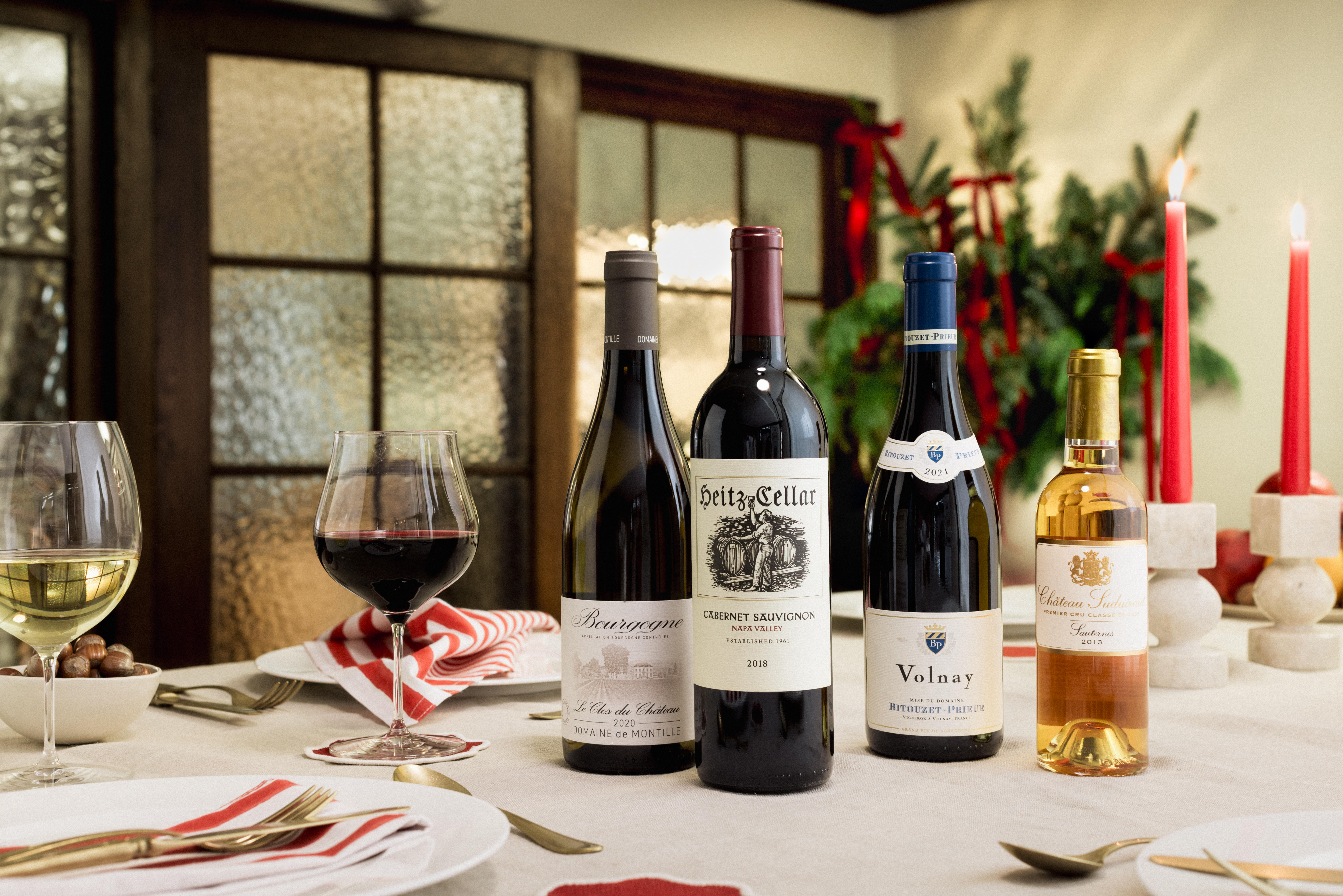 Four bottles of fine wine arranged on a festive dining table: Domaine de Montille Bourgogne 2020, Heitz Cellar Cabernet Sauvignon 2018, Volnay 2021, and Château Suduiraut Sauternes 2013.