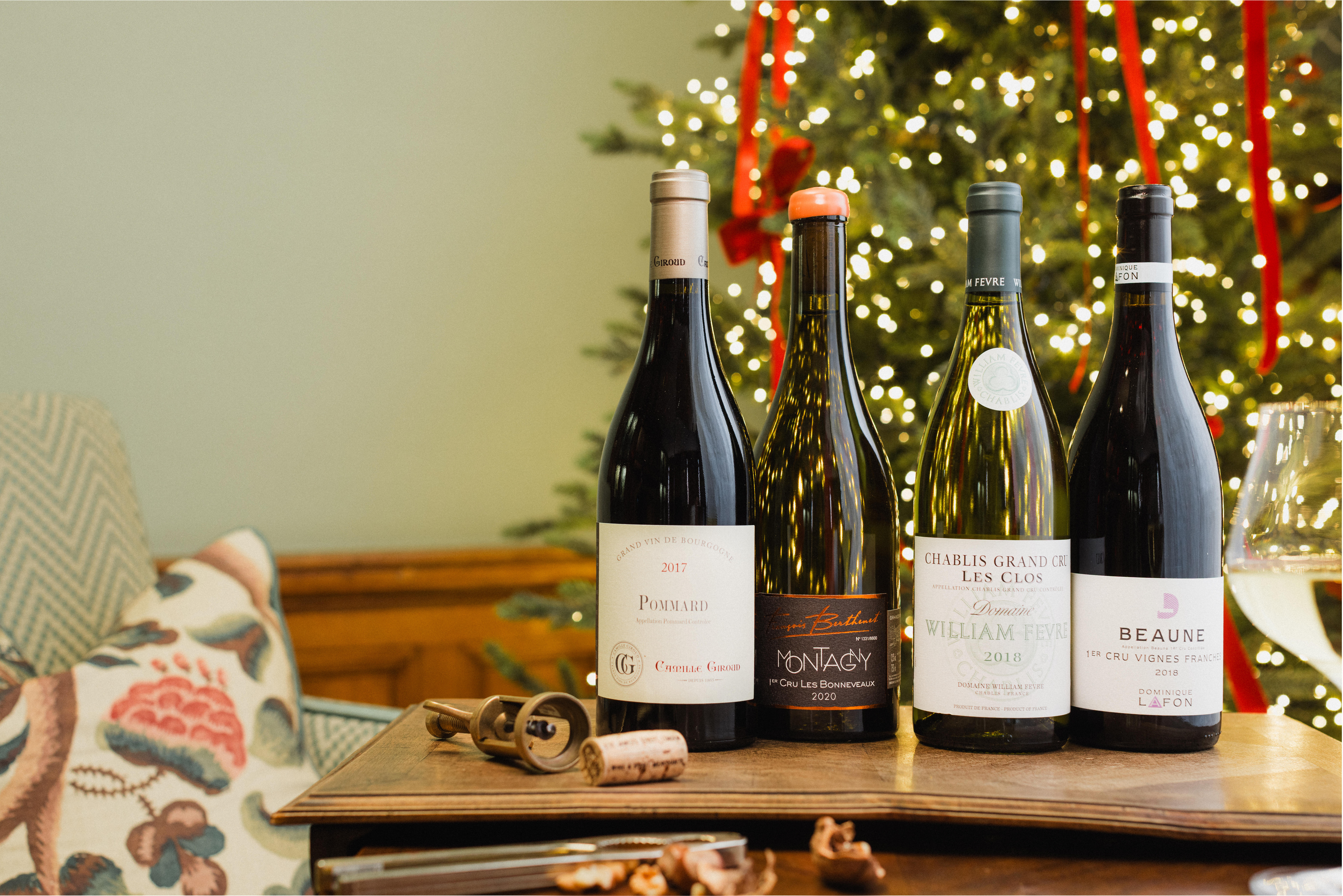 Four bottles of mature Burgundy wine on a festive table with a Christmas tree in the background, including Pommard, Montagny, Chablis Grand Cru, and Beaune.