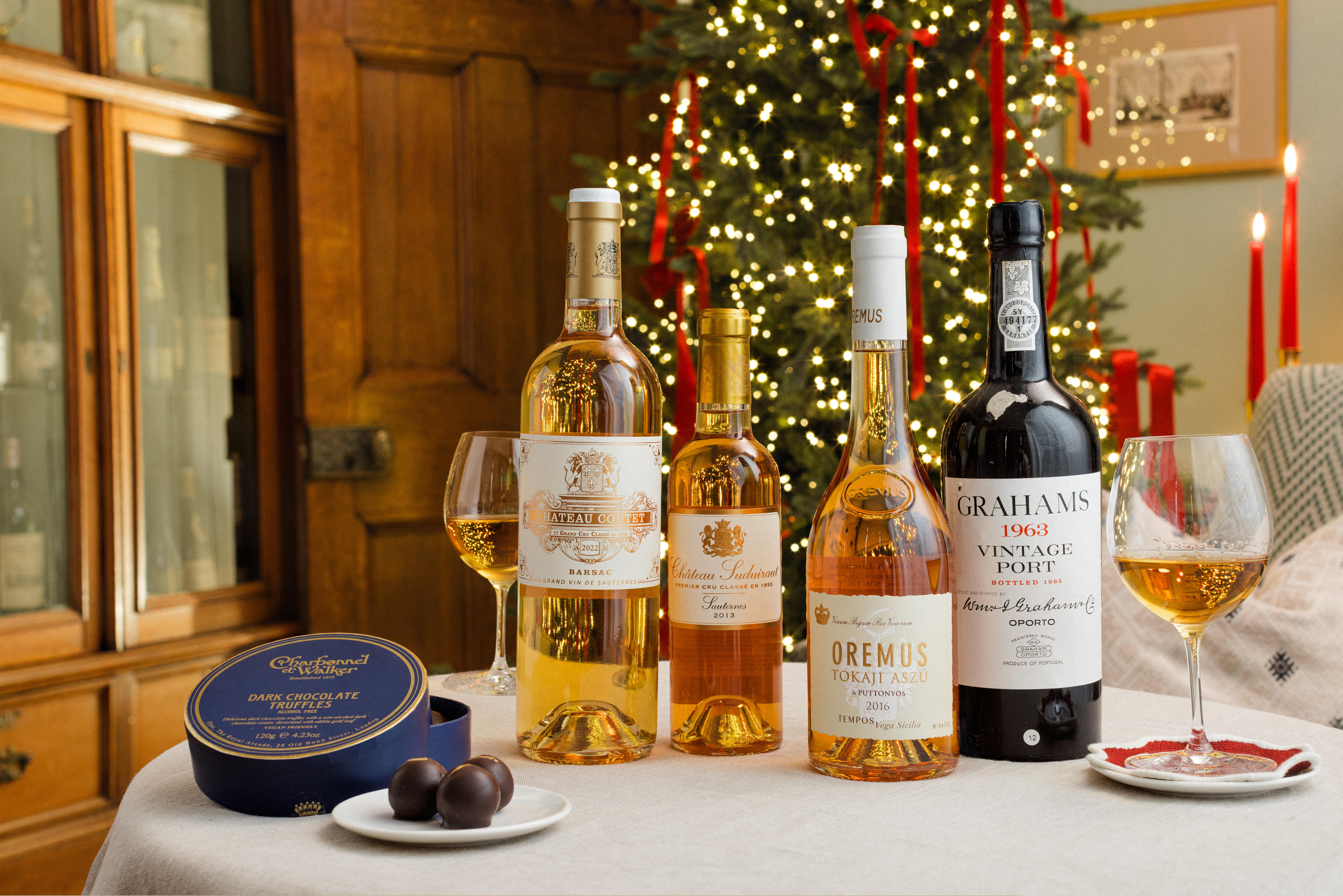 Christmas table with luxury Port and dessert wines, chocolate truffles, and festive decorations including a lit tree and candles.
