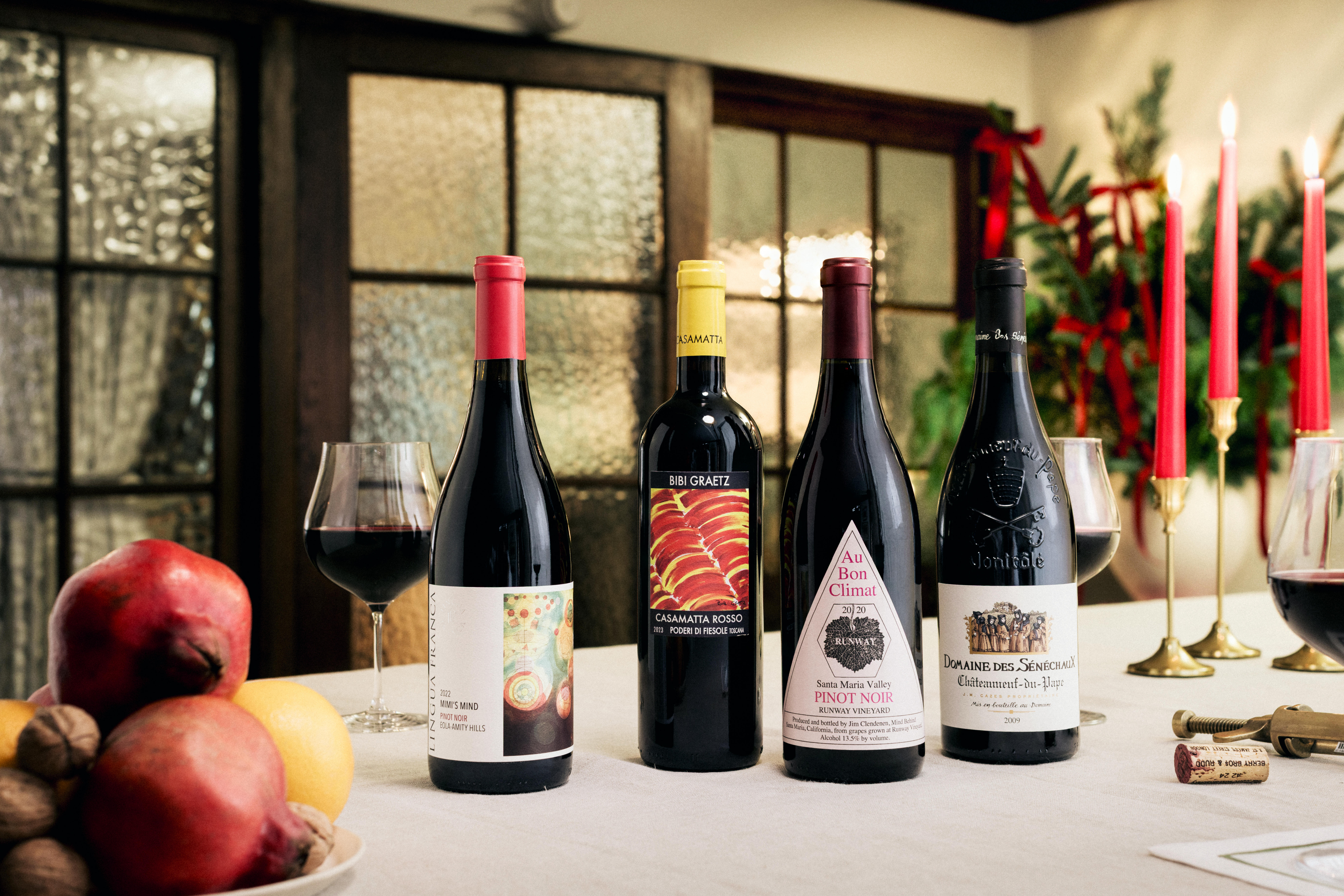 Four bottles of red wine displayed on a festive table with Christmas decorations, including Mind’s Mind Pinot Noir, Bibi Graetz Casamatta Rosso, Au Bon Climat Pinot Noir, and Domaine des Sénéchaux Châteauneuf-du-Pape.