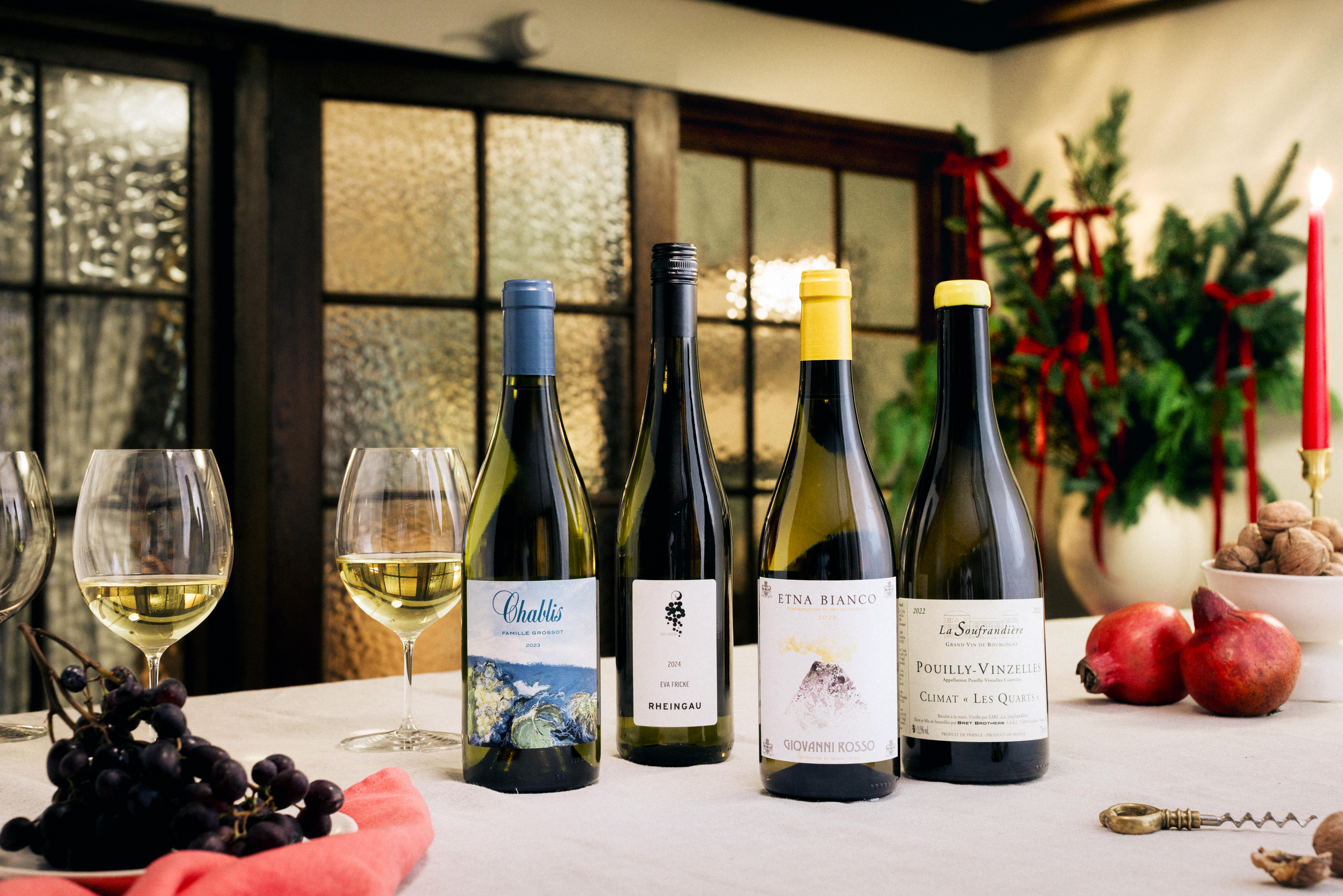 Four bottles of white wine displayed on a festive table with Christmas decorations, including Chablis, Rheingau, Etna Bianco, and Pouilly-Vinzelles. Two glasses of white wine, grapes, pomegranates, and a candle add to the seasonal setting.