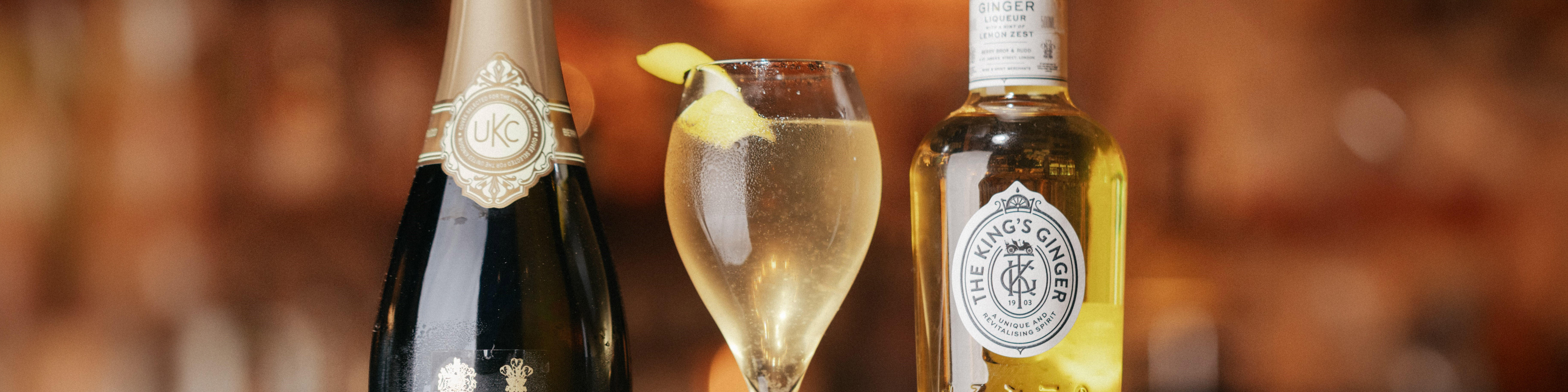 A glass of a golden cocktail, garnished with lemon peel, served between a bottle of The King’s Ginger liqueur and a bottle of sparkling wine, set against a warm, amber background.