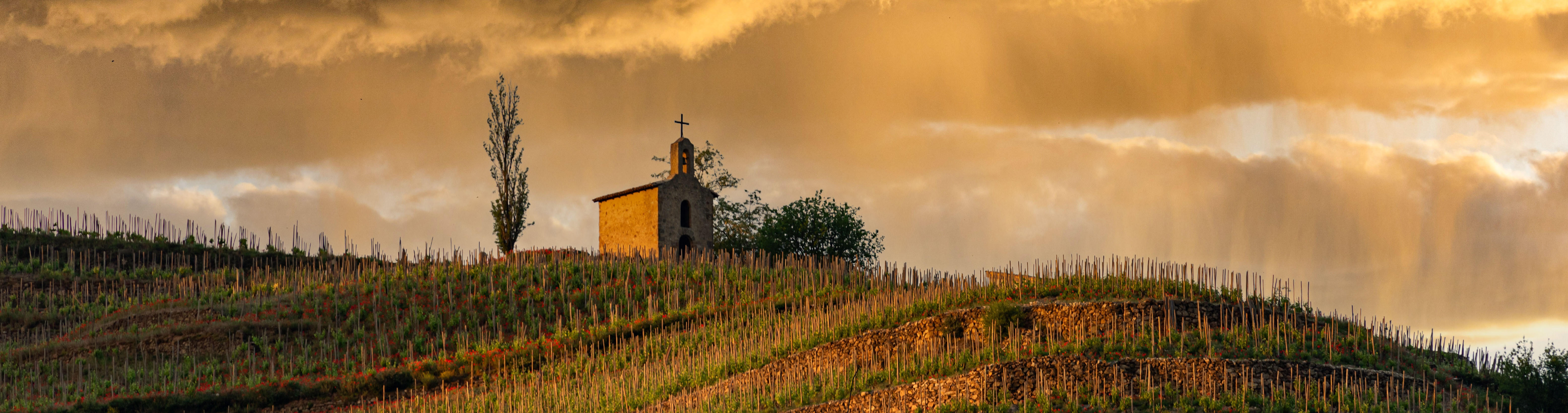The historic chapel of Hermitage perched on a terraced hillside vineyard in the Rhône Valley, under dramatic golden clouds at sunset.