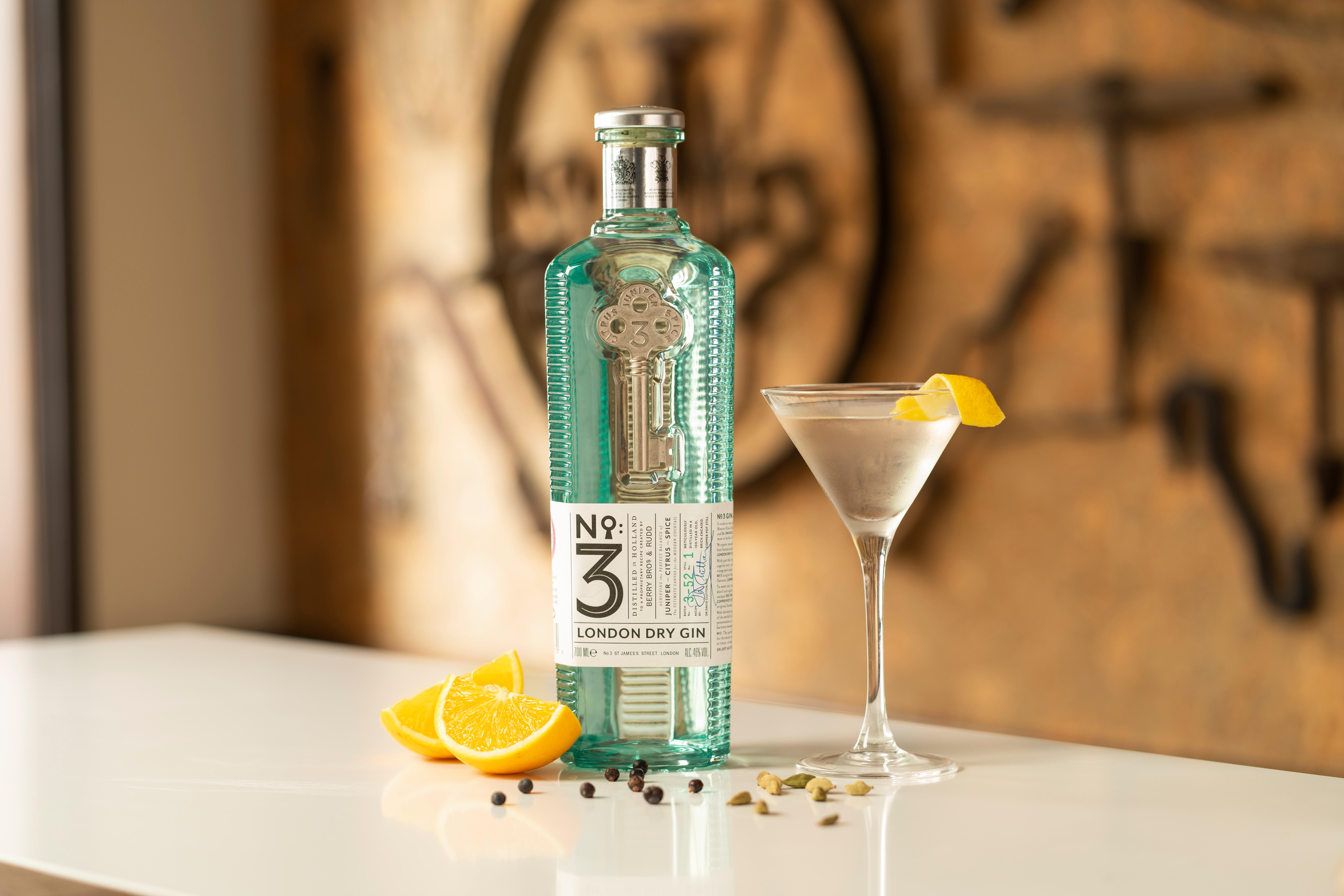A bottle of No.3 London Dry Gin standing on a white surface beside a Martini glass garnished with a lemon twist. Orange slices, juniper berries, and cardamom pods are scattered in the foreground.