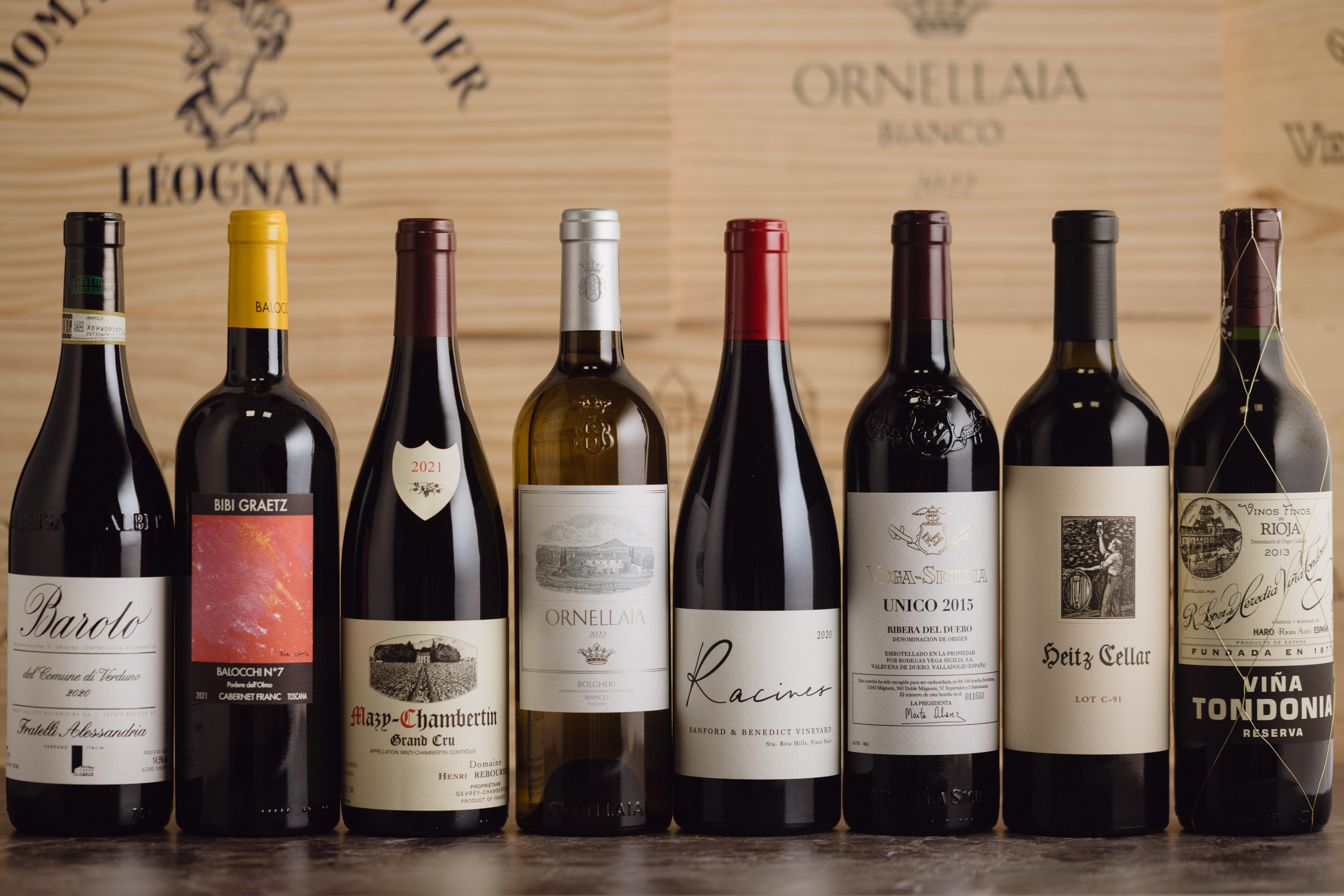A selection of fine wines displayed with wooden crates, featuring Barolo, Cabernet Franc, Mazis-Chambertin, Ornellaia Bianco, Pinot Noir, Vega-Sicilia, Heitz, and Rioja Reserva.
