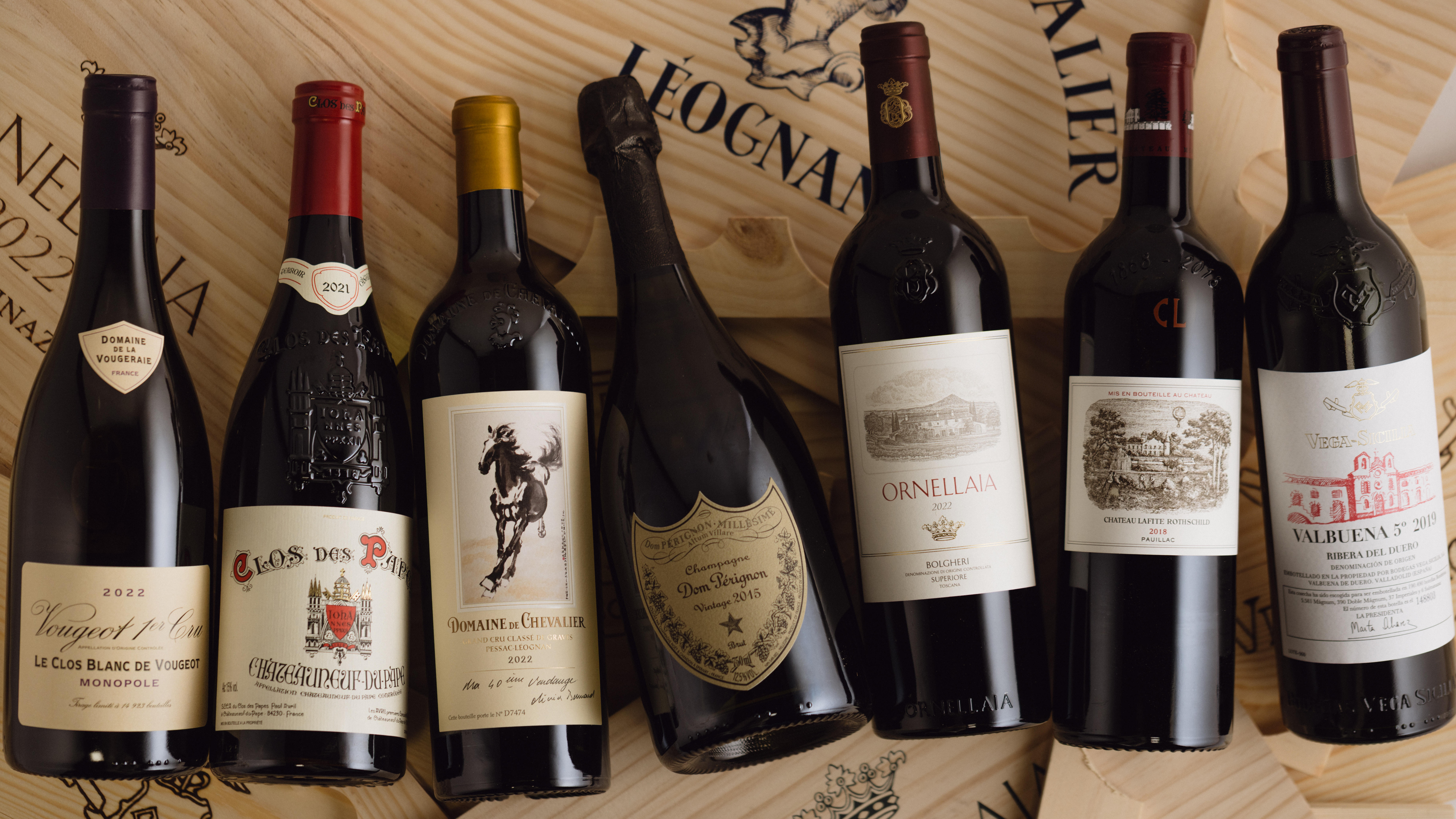 A selection of seven fine wine bottles arranged on wooden wine cases stamped with prestigious vineyard names. The bottles include a mix of red and sparkling wines from renowned producers.