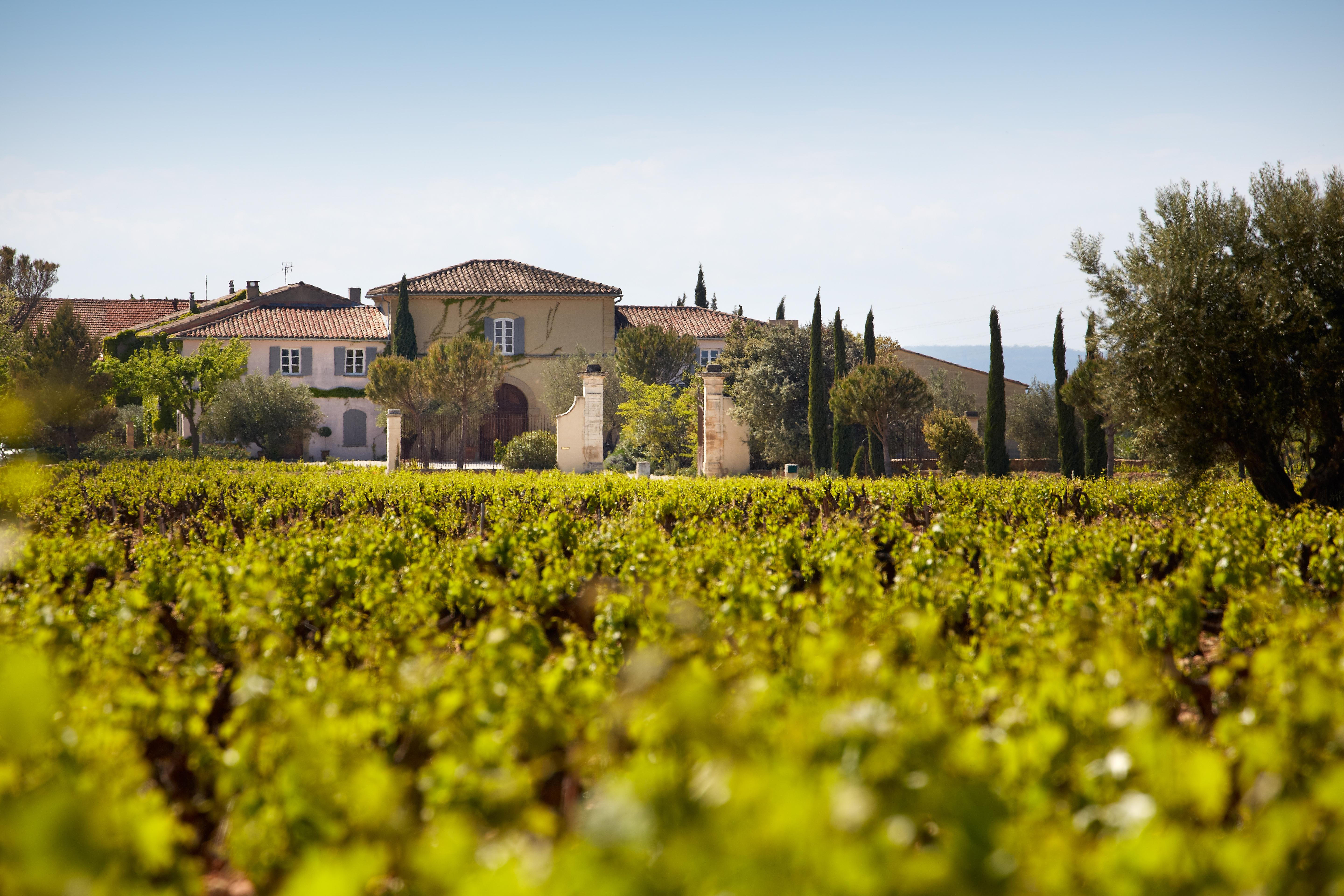 Château de Beaucastel estate surrounded by lush green vineyards under a bright blue sky, with traditional stone buildings and terracotta roofs framed by tall cypress trees.