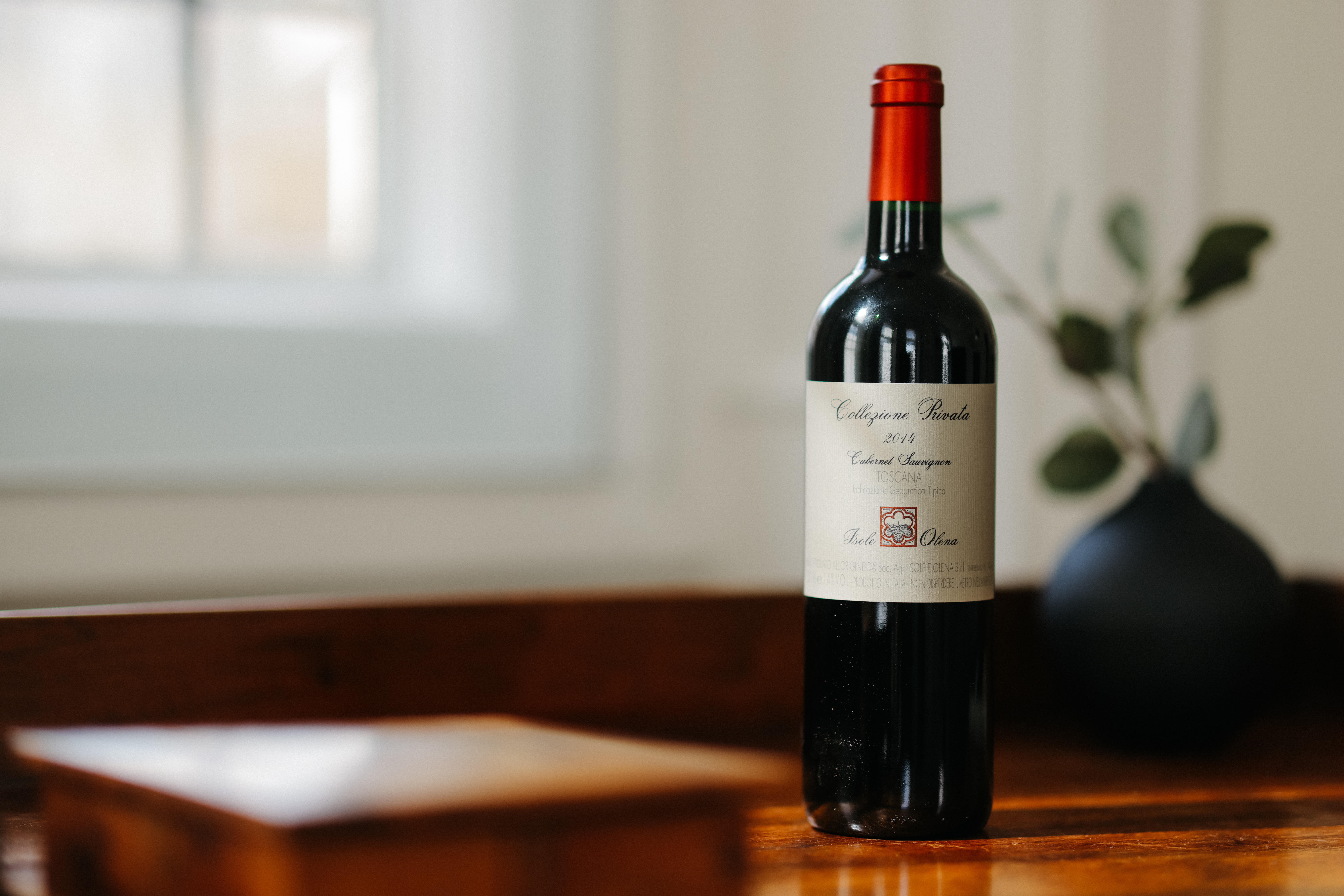 A bottle of Isole e Olena Collezione Privata Cabernet Sauvignon 2014 on a wooden surface, with a blurred vase and window in the background.