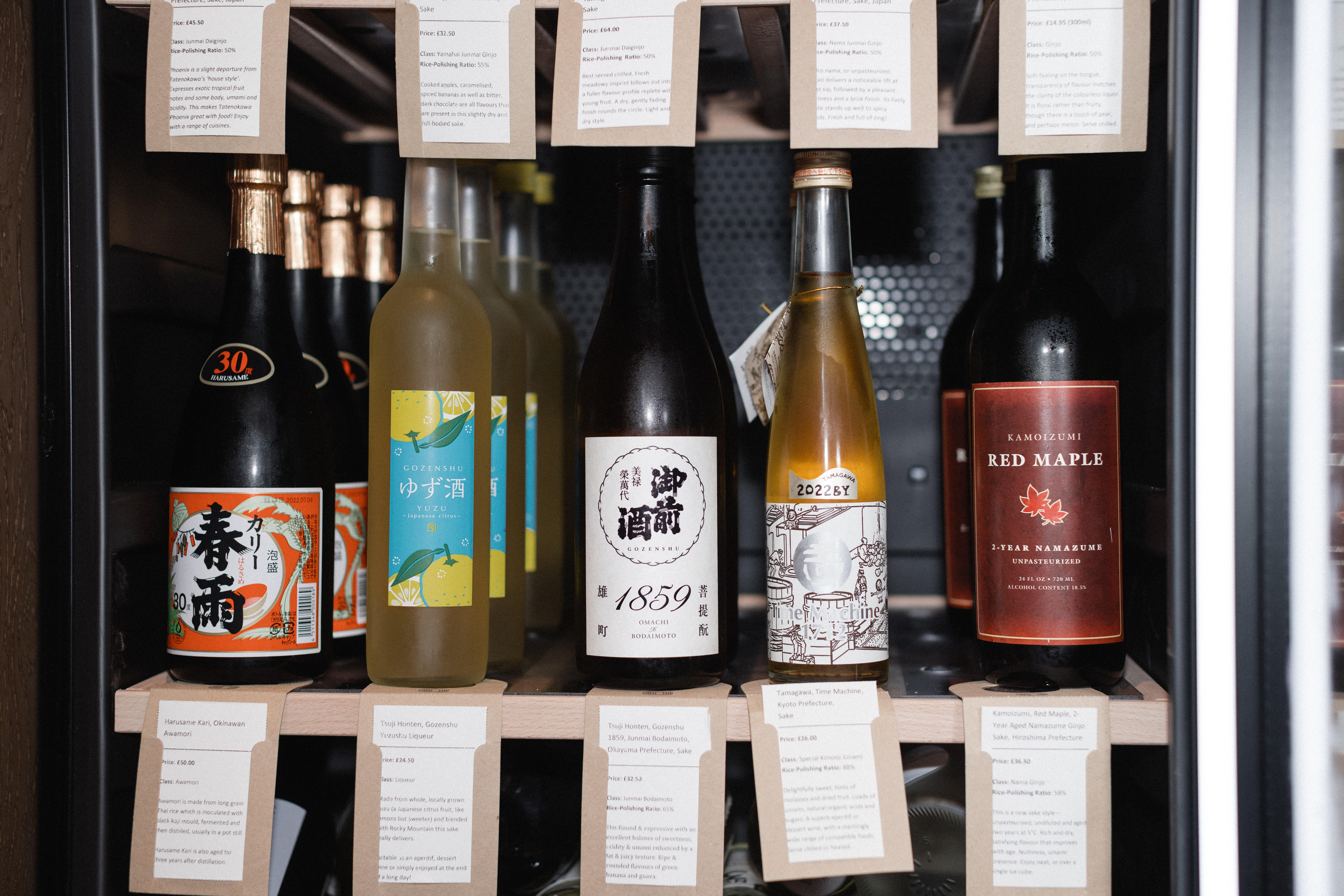 A selection of sake bottles displayed in a refrigerated cabinet, featuring various labels and styles, including traditional Japanese designs and modern branding. Visible bottles include sake with vibrant labels, a yuzu-infused sake, and premium options.