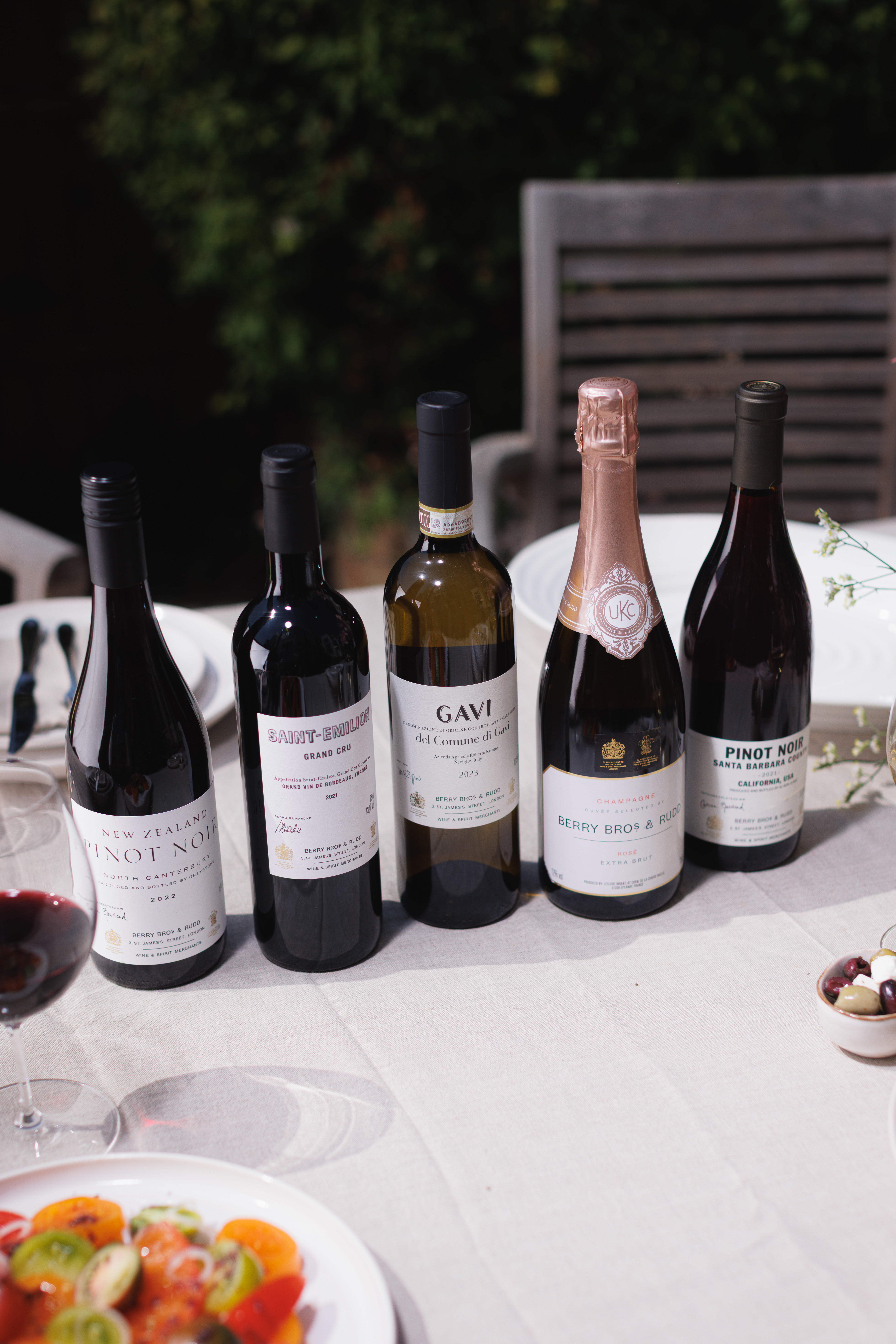 A selection of five wines from Berry Bros. & Rudd’s Own Selection displayed on a dining table, including New Zealand Pinot Noir, Saint-Emilion Grand Cru 2021, Gavi del Comune di Gavi 2023, Champagne Rosé Brut, and California Pinot Noir.