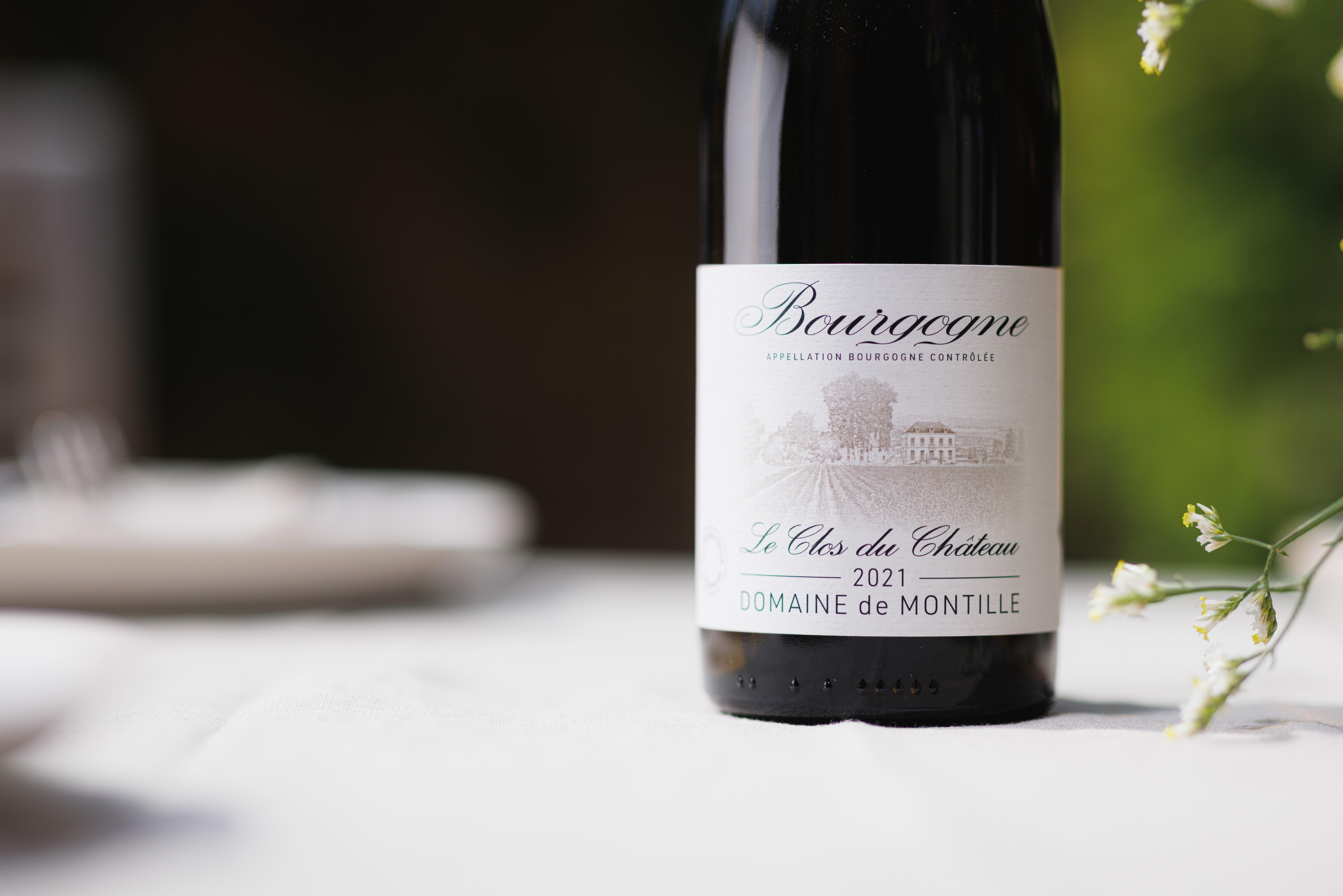 A bottle of Bourgogne Le Clos du Château 2021 from Domaine de Montille, featuring an illustrated vineyard and château on the label, set against a softly blurred dining background with white flowers.