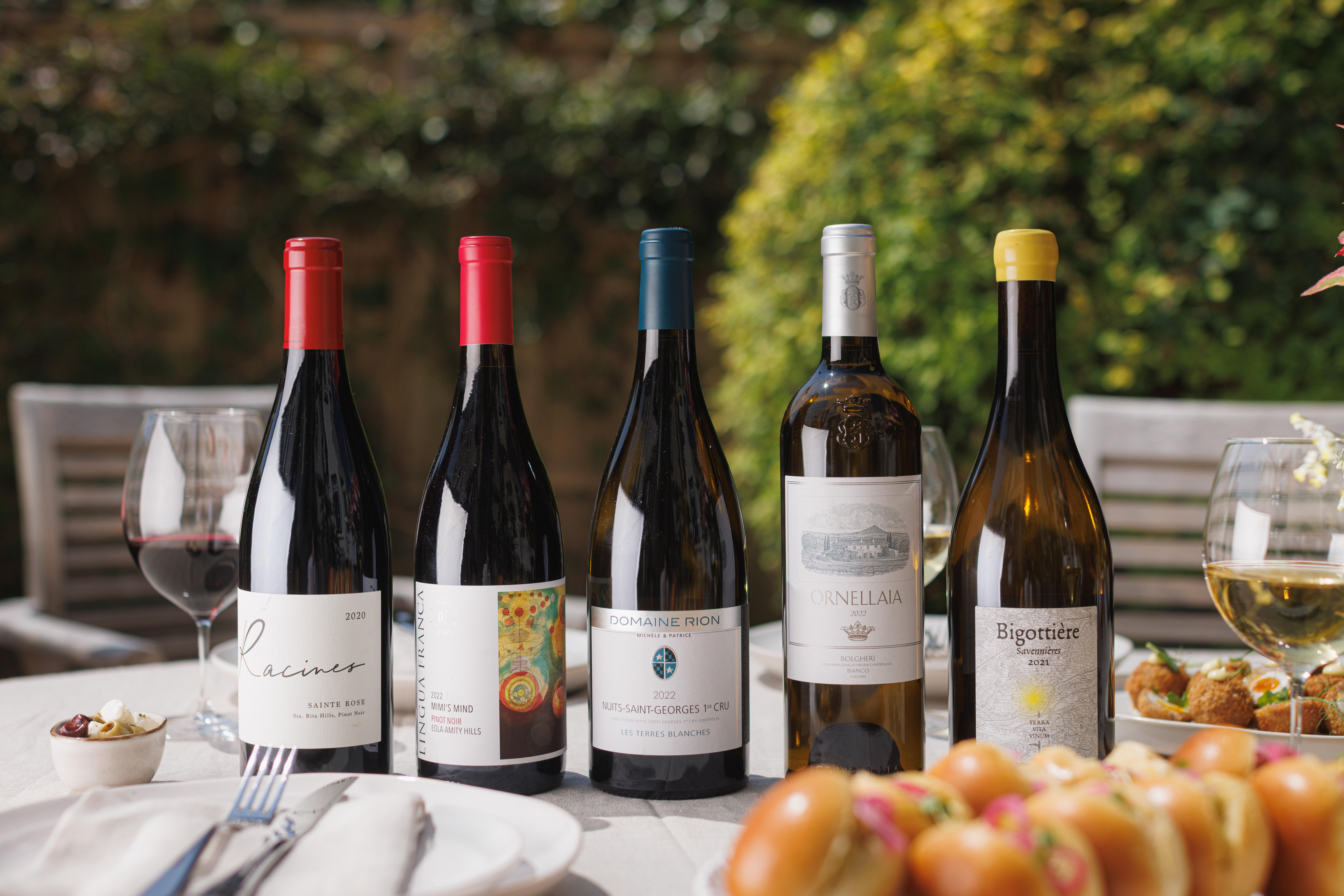 Five wine bottles with red and white wine glasses, plates of appetizers, and a casual outdoor dining setup.