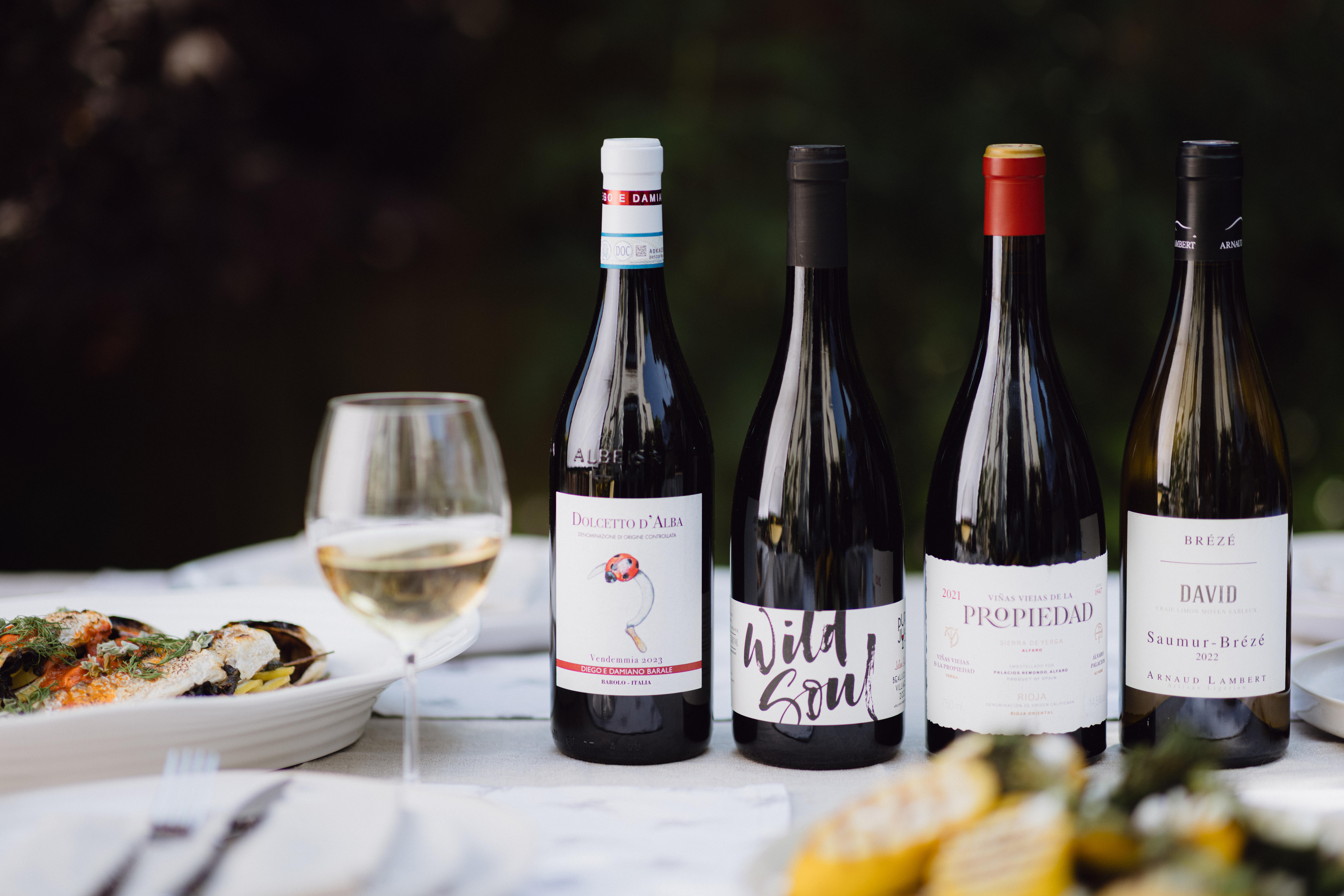 A selection of new arrival wines on a summer table setting, featuring four bottles and a glass of white wine alongside grilled seafood and vegetables.