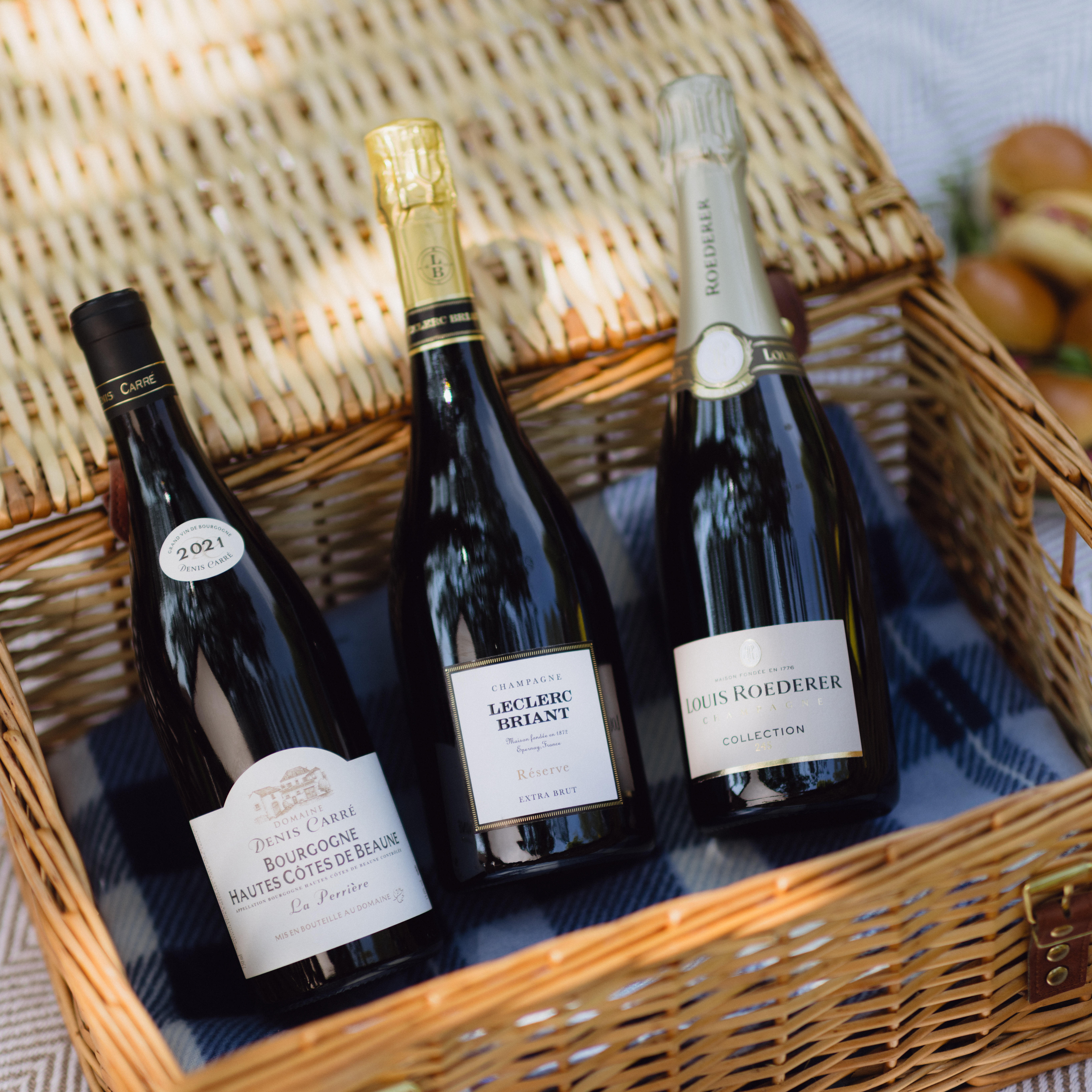A trio of fine wines nestled in a picnic hamper on a blanket, featuring Bourgogne Hautes Côtes de Beaune, Leclerc Briant Champagne, and Louis Roederer Collection — part of Berry Bros. & Rudd’s mixed case selection.