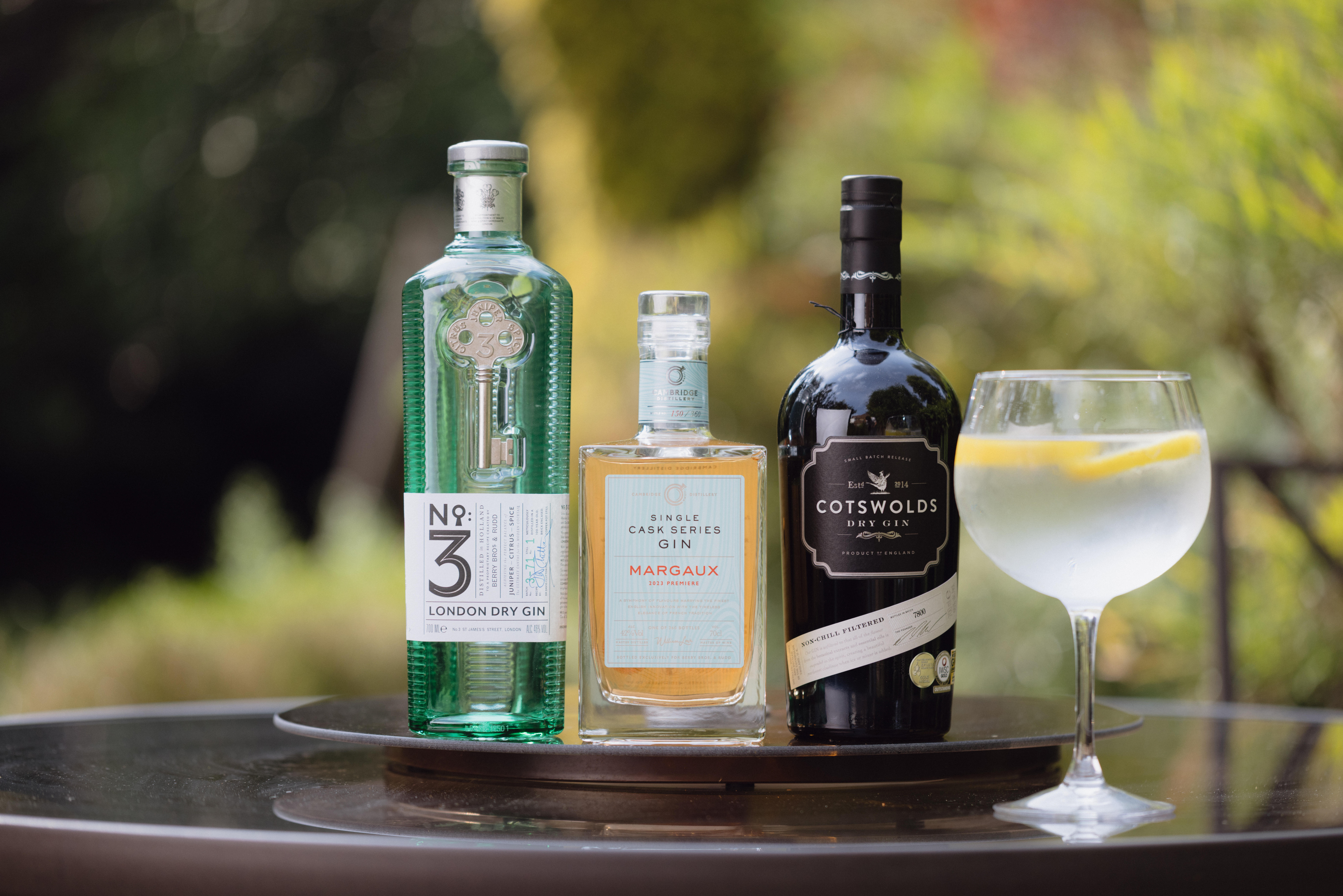 Three bottles of premium gin—No.3 London Dry Gin, Single Cask Series Gin Margaux, and Cotswolds Dry Gin—displayed on an outdoor table beside a gin and tonic cocktail garnished with lemon slices.