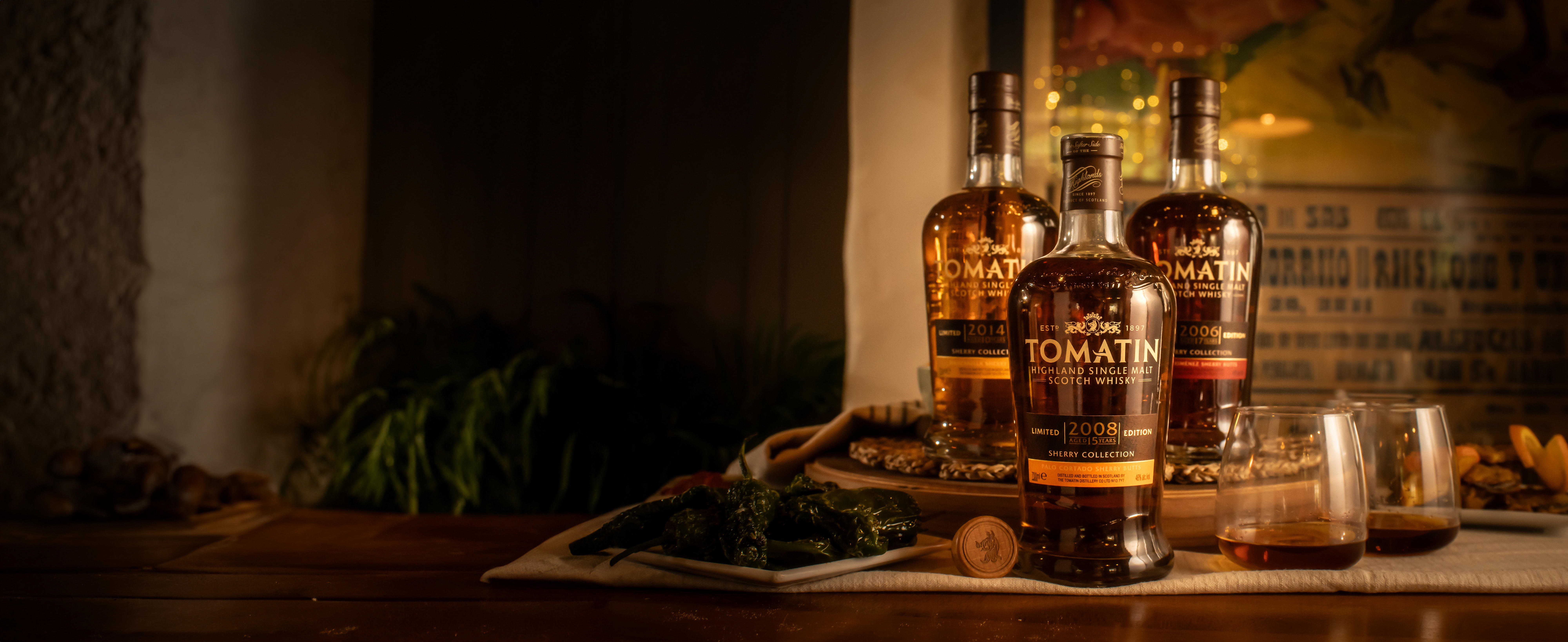 Three bottles of Tomatin Highland Single Malt Scotch Whisky displayed on a wooden table with two whisky glasses and a cork in the foreground, warm ambient lighting and a vintage poster in the background.