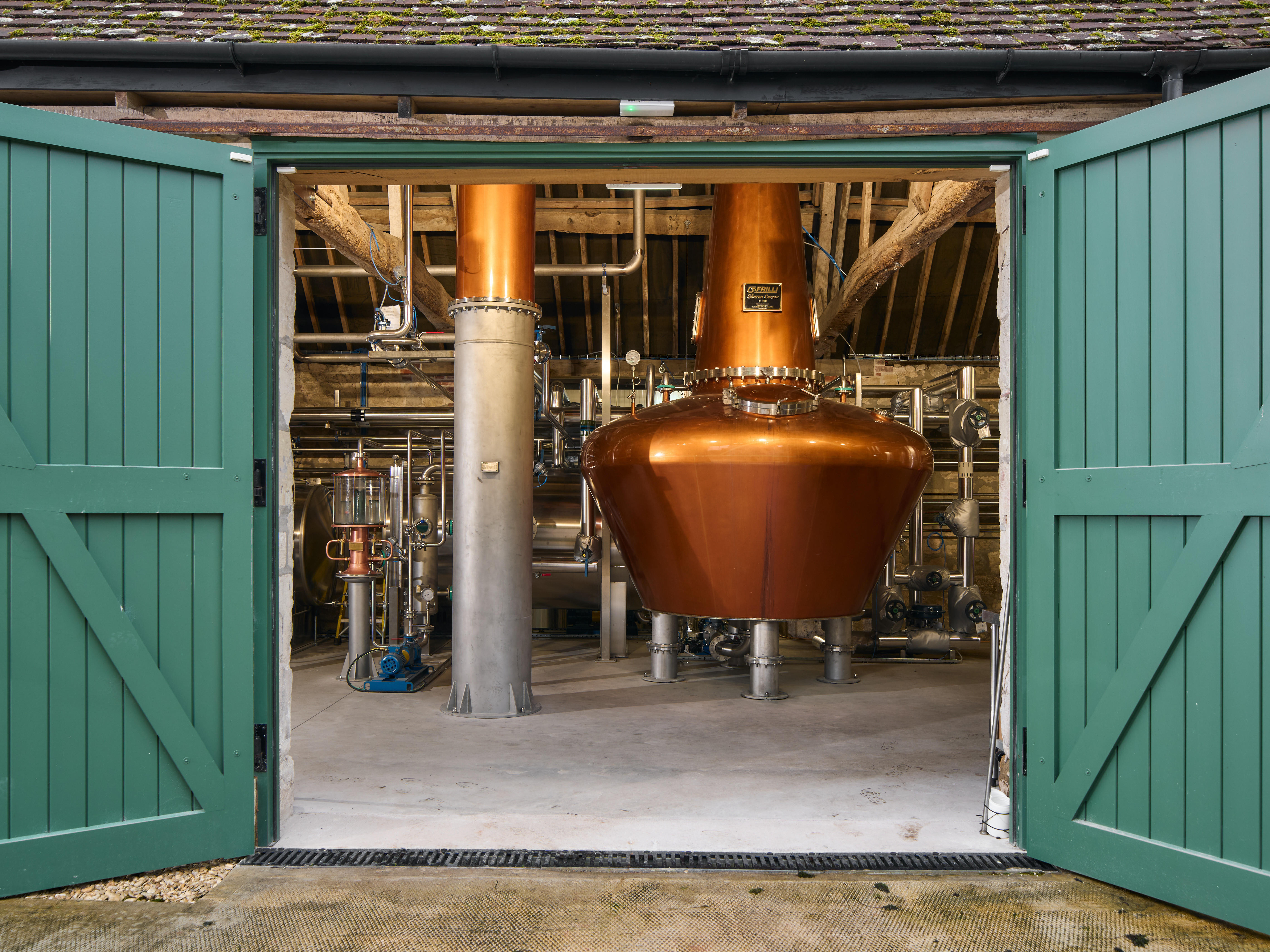 Copper pot still inside Witchmark Distillery, viewed through open green double doors, with stainless steel pipes and wooden beams visible in the background.