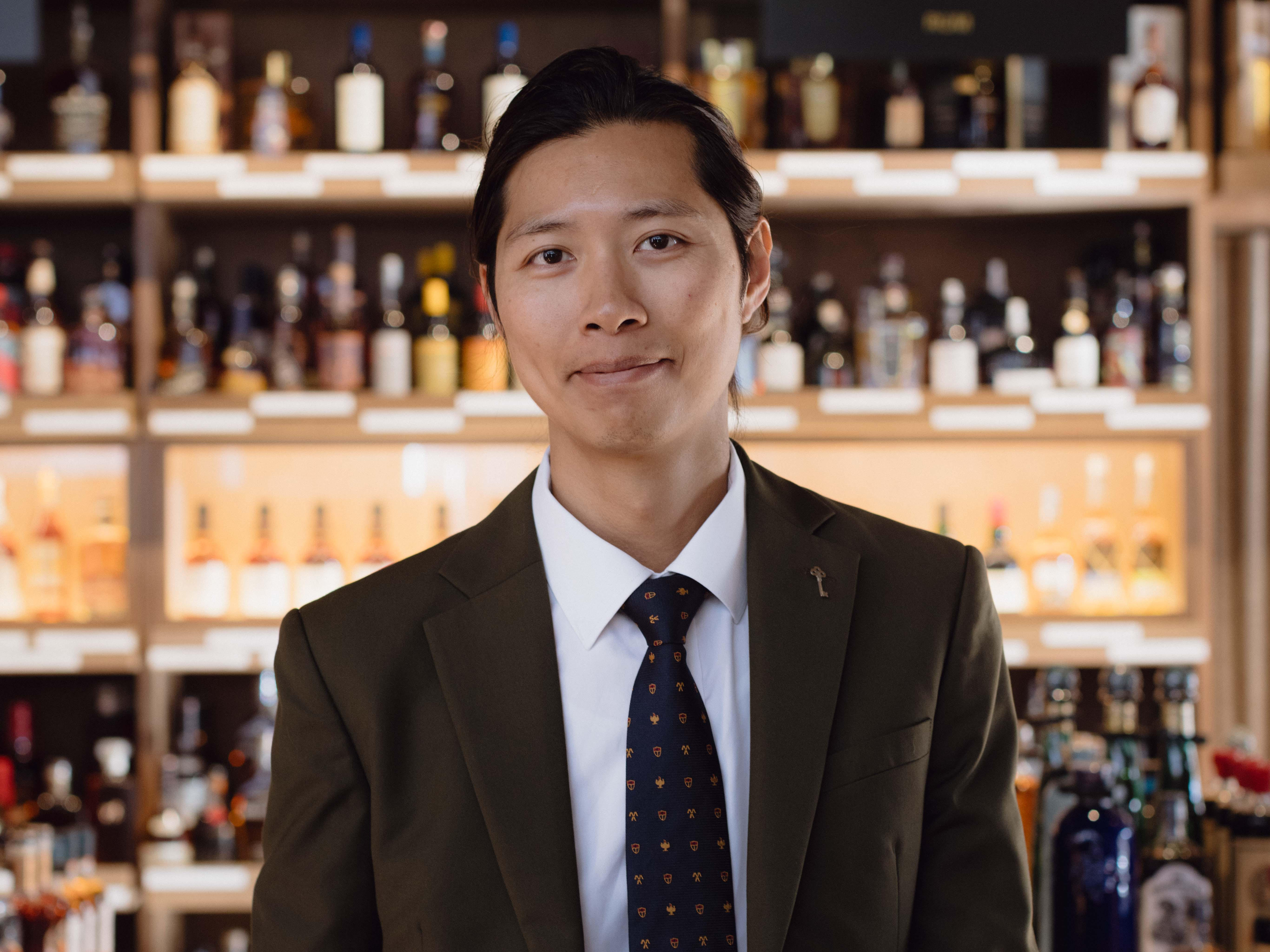 Shop manager wearing a dark green suit and patterned tie, standing in front of shelves filled with various bottles of spirits, including whiskies, rums, and other liquors.