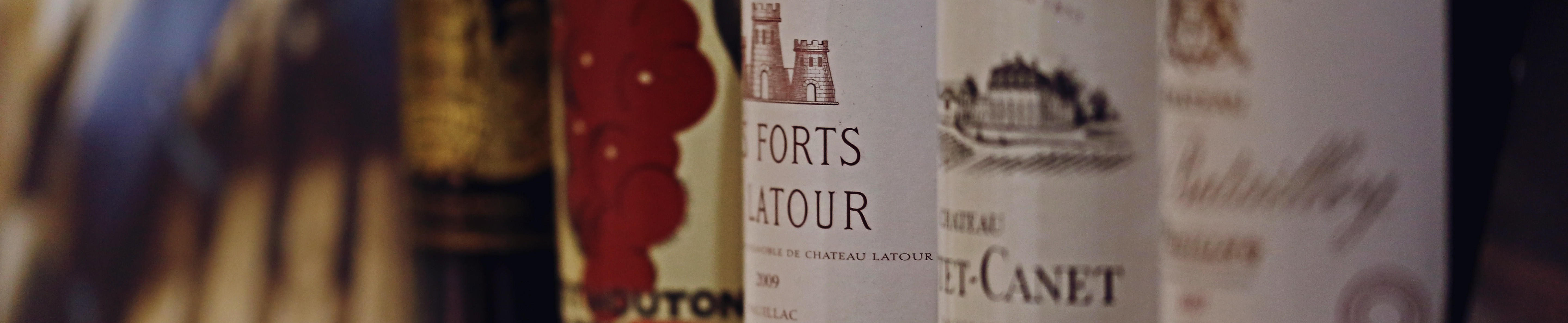 A close-up of fine Bordeaux wine labels, including Les Forts de Latour and Château Mouton Rothschild, with soft focus on surrounding bottles.