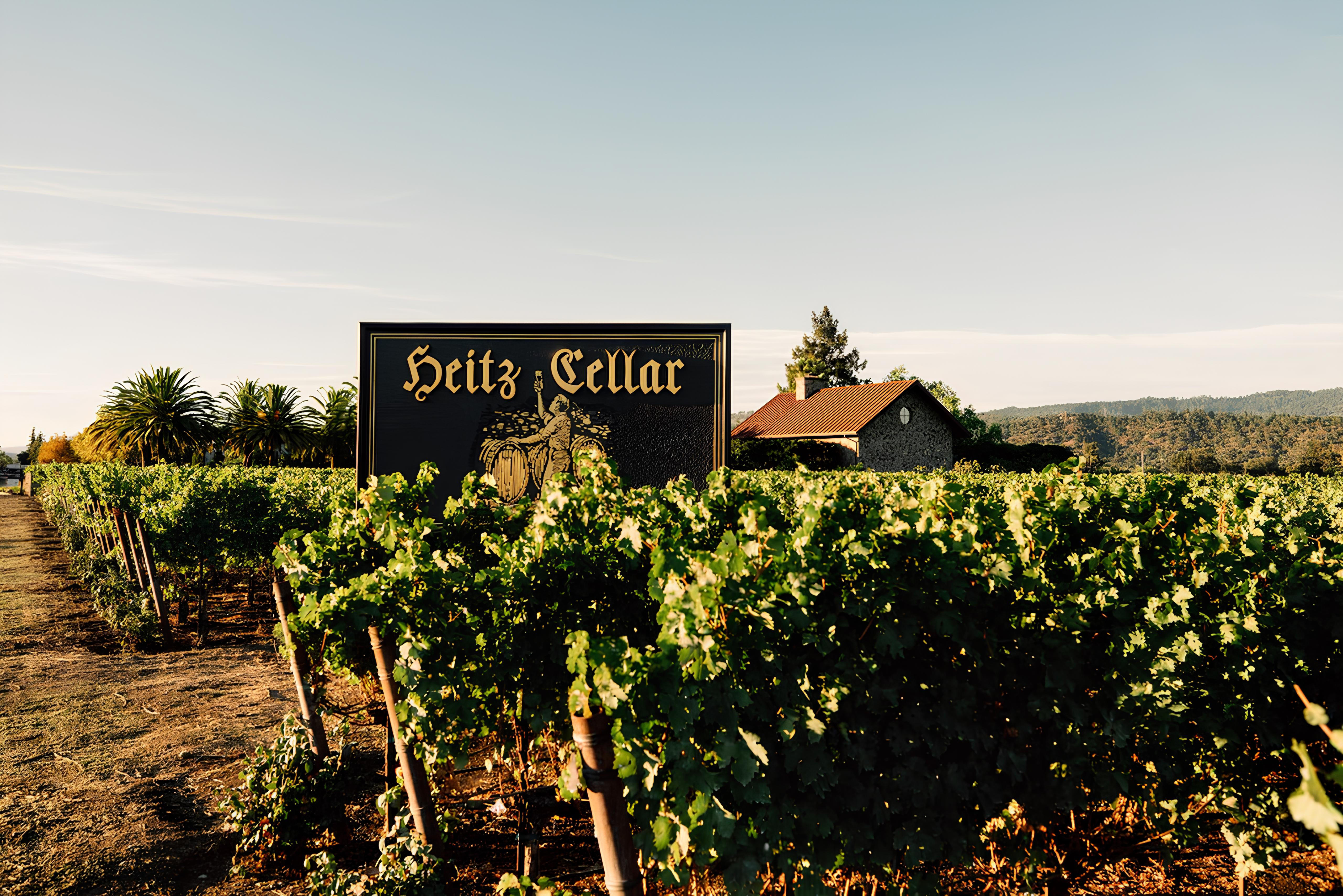 Heitz Cellar vineyard in Napa Valley with lush green grapevines under clear skies, featuring a prominent black and gold Heitz Cellar sign and a stone building with a red-tiled roof in the background.