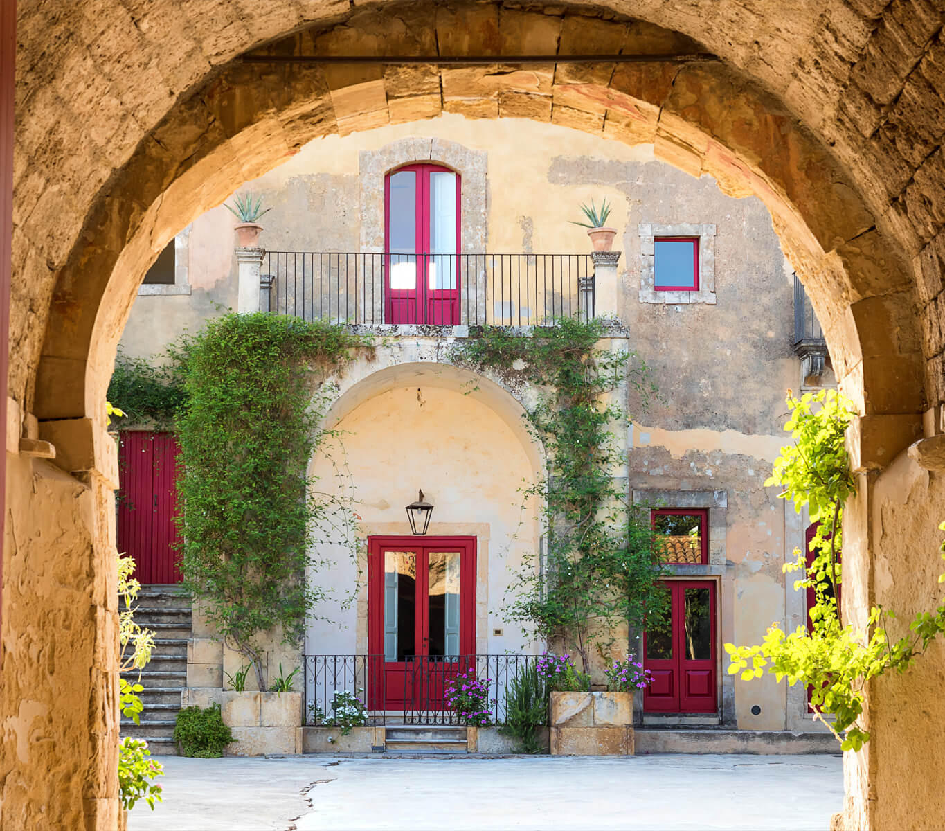Red doors with wrought-iron balconies overlooking a sunlit Tuscan vineyard.