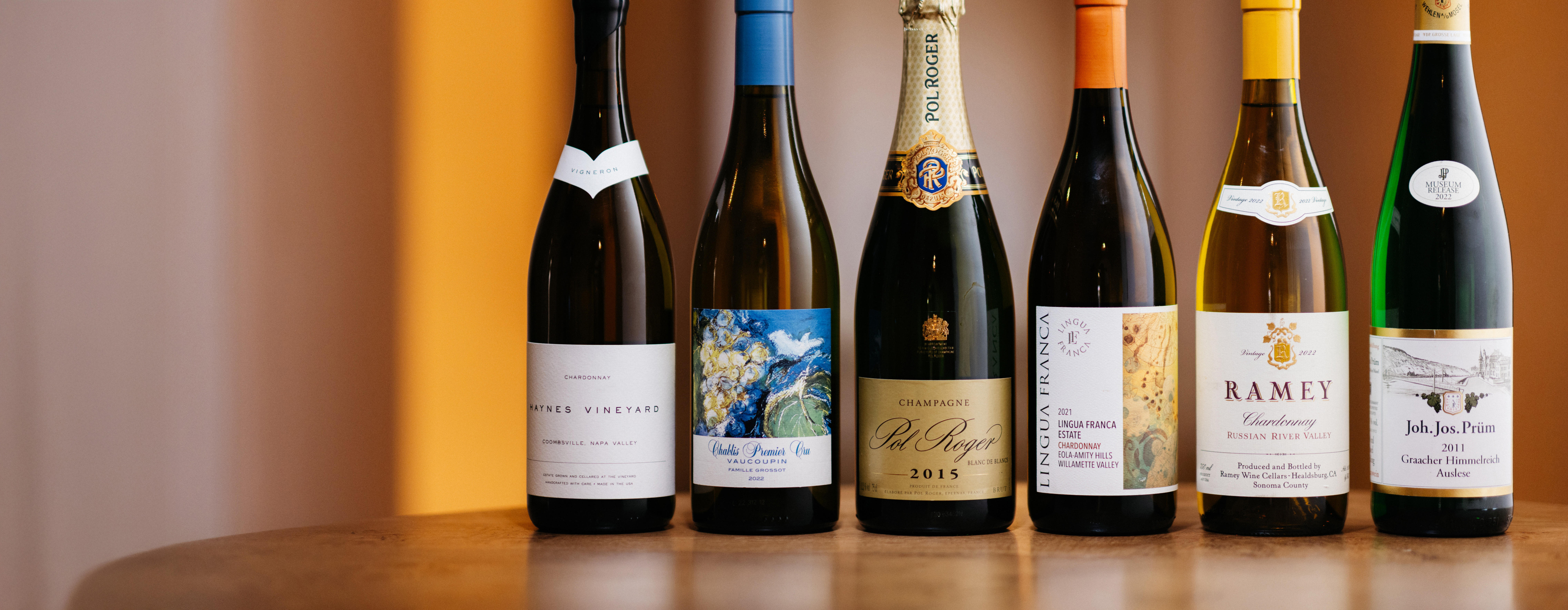 Six assorted wine bottles on a wooden surface, featuring Chardonnay, Pinot Noir, Champagne, and Riesling from renowned regions like Napa Valley, Burgundy, and Mosel.